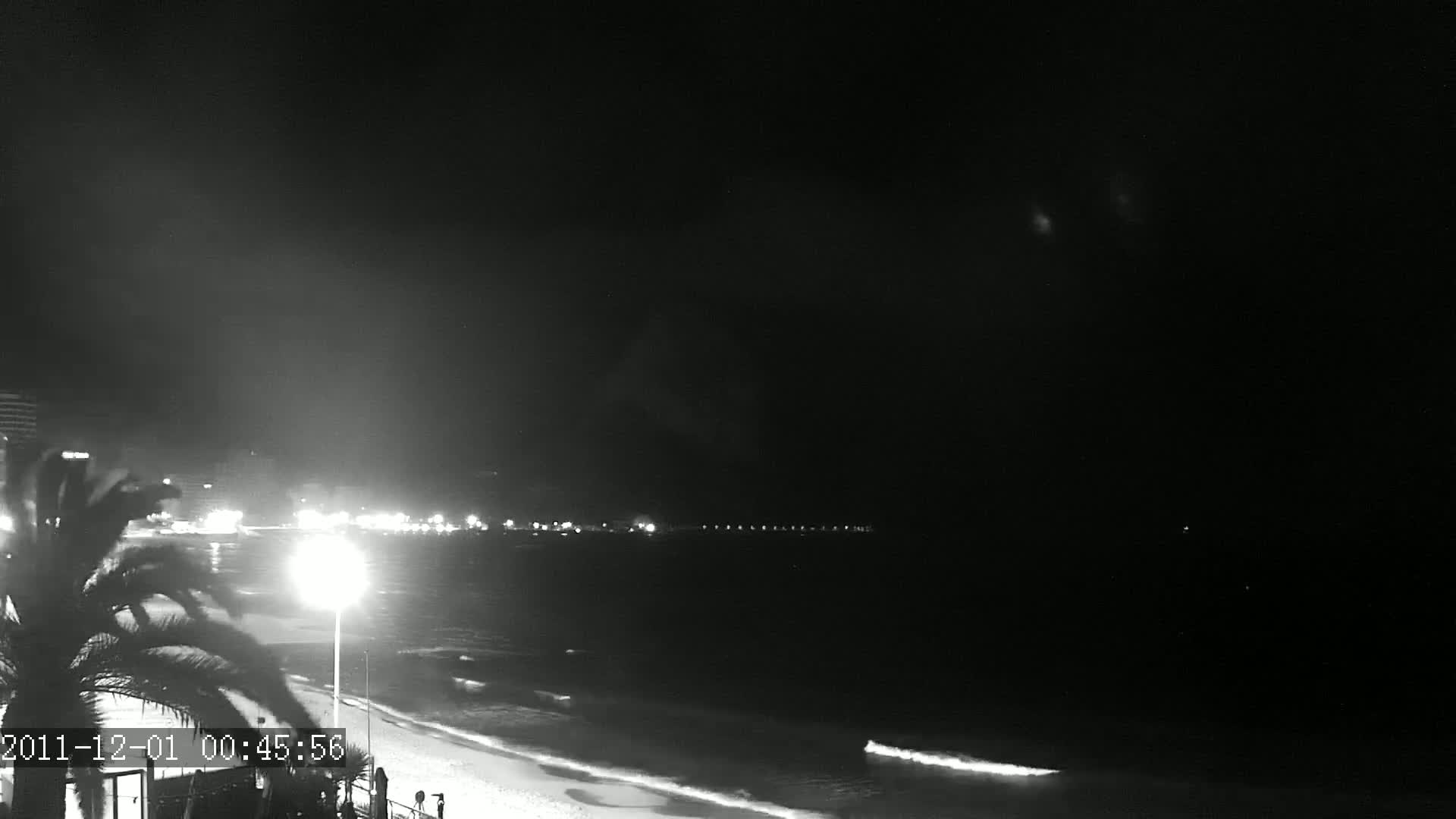 A nighttime beach scene shows gently lapping waves on a sandy shore, with lights from a distant city illuminating the dark sky.