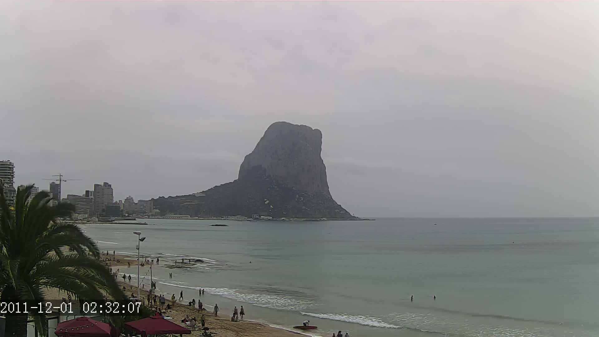 A large, dark gray rock formation rises from the sea under an overcast sky, as people relax on a sandy beach in front of a cityscape.