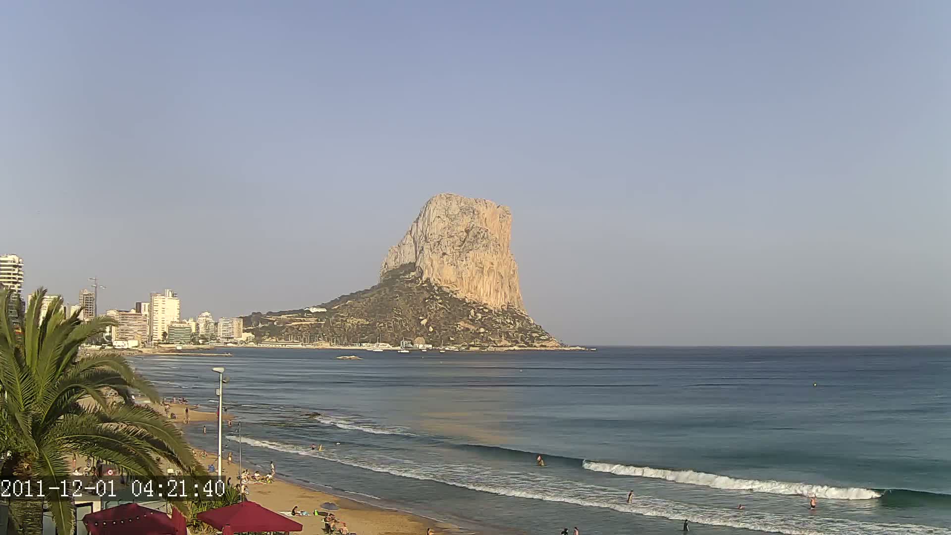 A sandy beach with calm waves and several people swimming in the ocean, overlooks a city skyline with a large, tan, rocky mountain in the background under a clear sky.