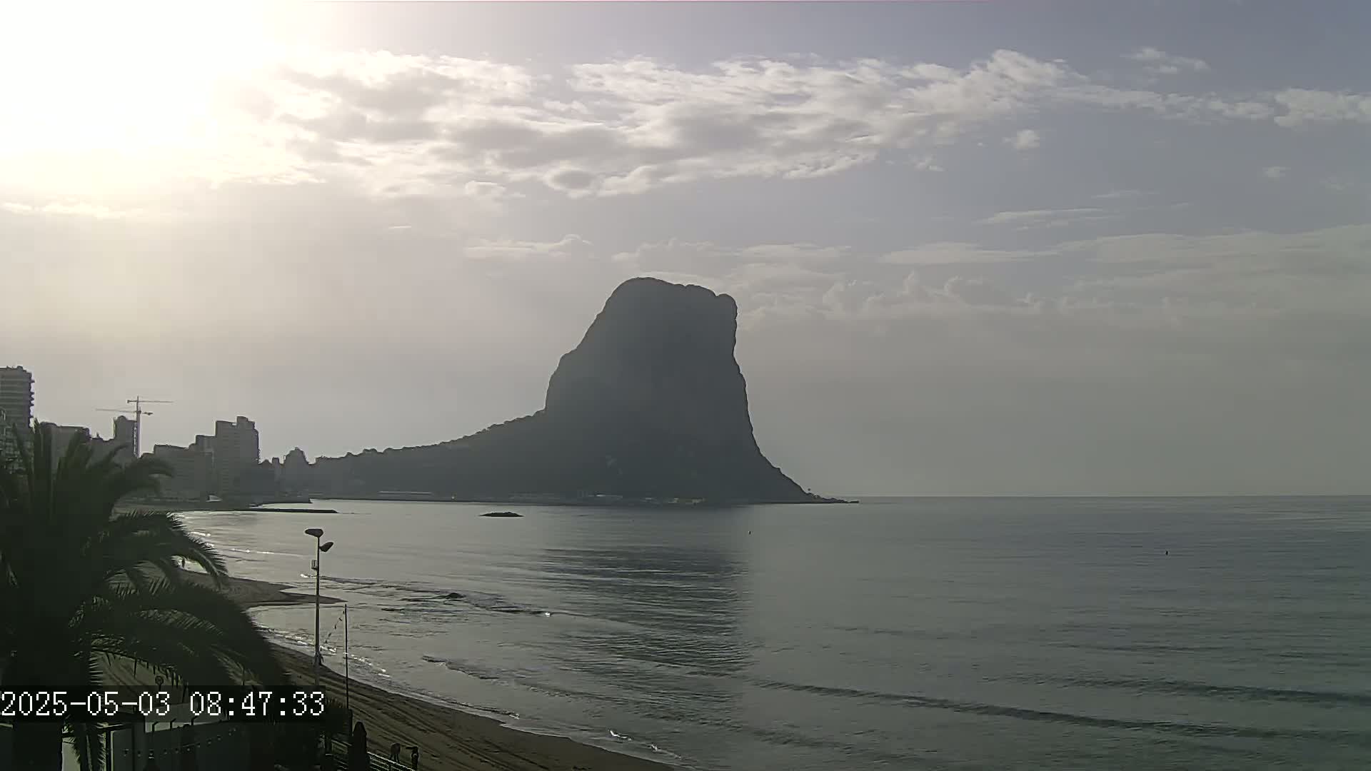 A large, dark mountain rises from the calm sea under a mostly cloudy sky, with a sandy beach and buildings visible in the foreground.