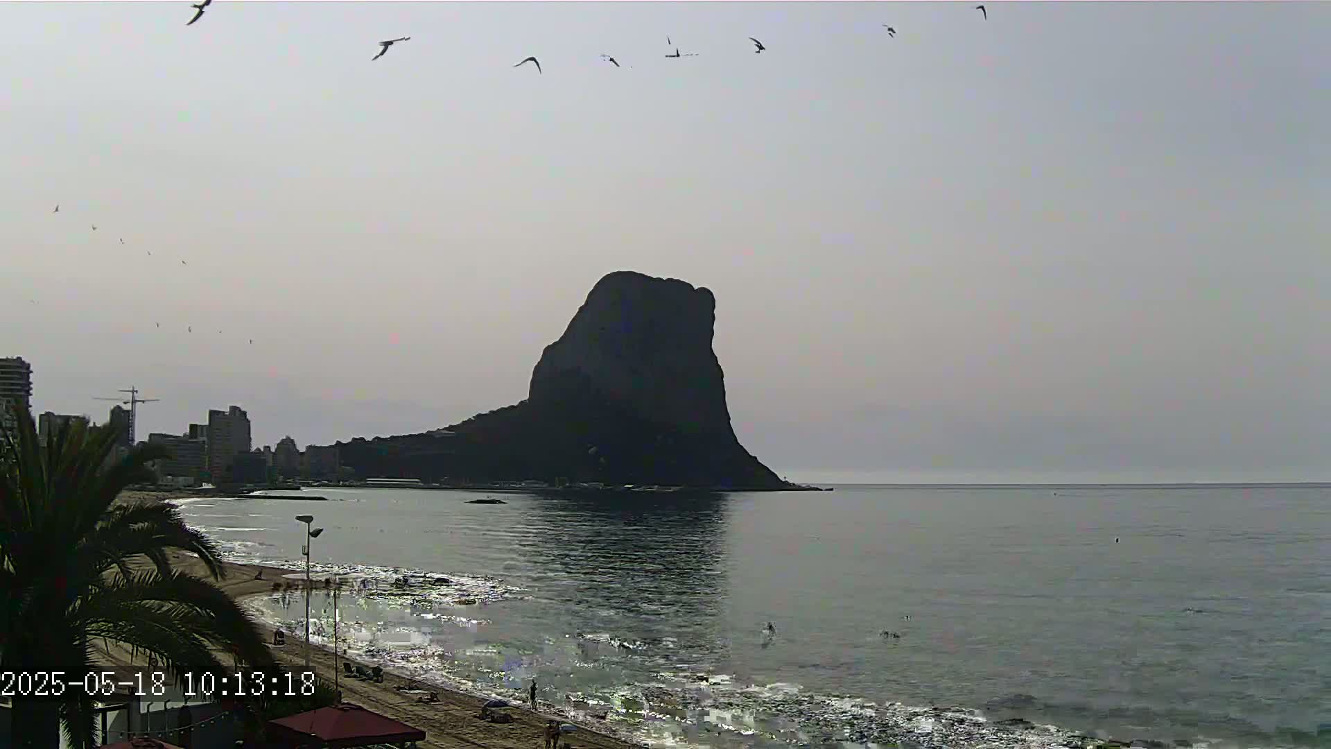 A large, dark rock formation rises from the calm, gray sea under a hazy sky, with birds flying overhead and a sandy beach with people visible in the foreground.