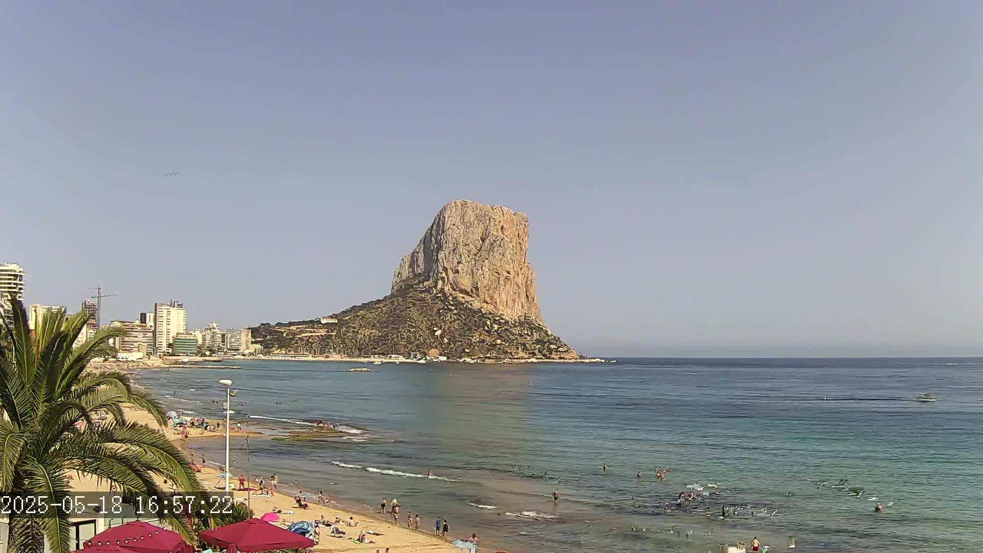 A sandy beach with swimmers in the calm ocean,  under a clear sky, features a large, rocky, tan-colored island in the distance.