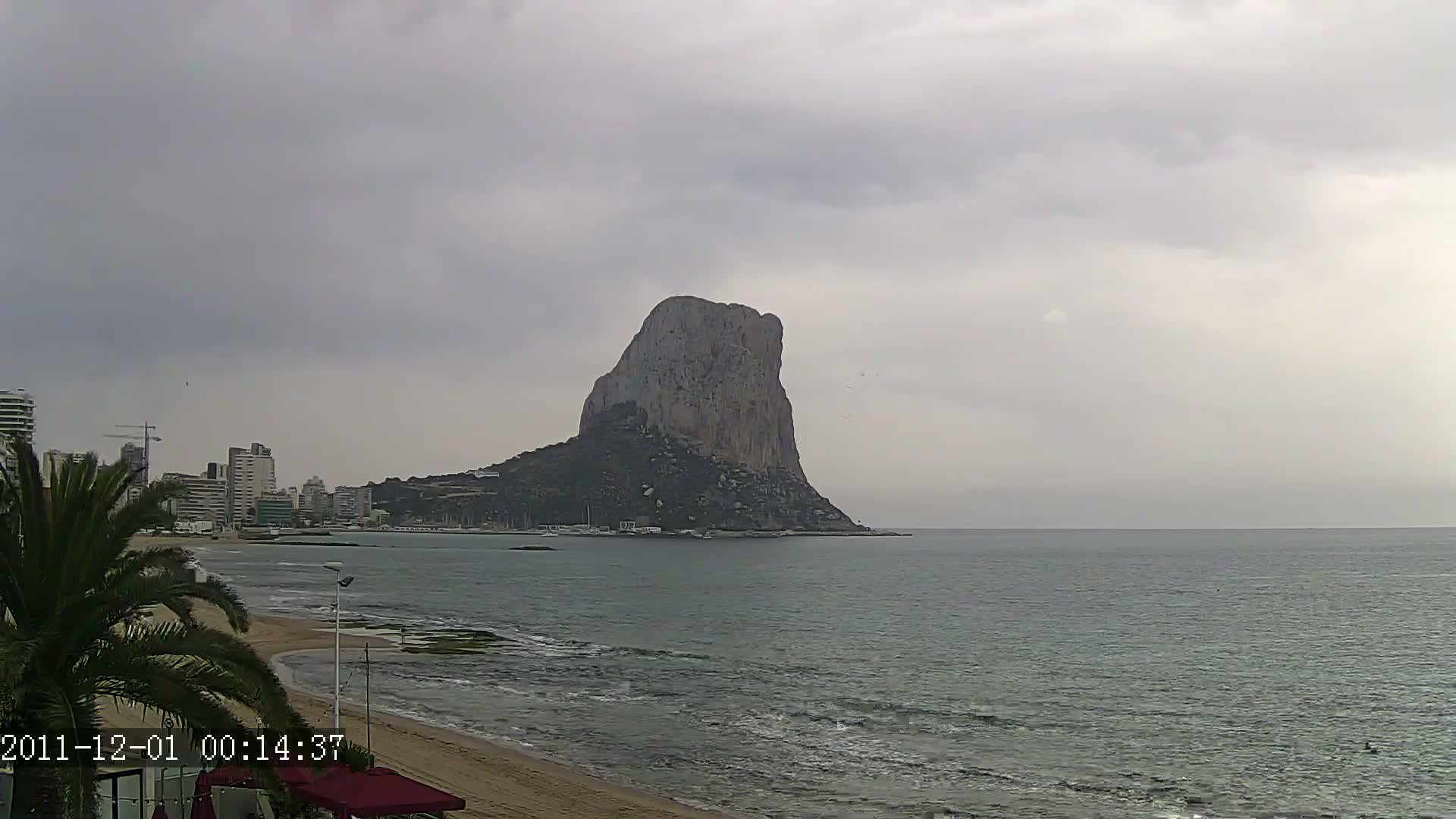 A large, rocky island stands in a calm sea under an overcast sky, with a sandy beach and buildings visible in the background.