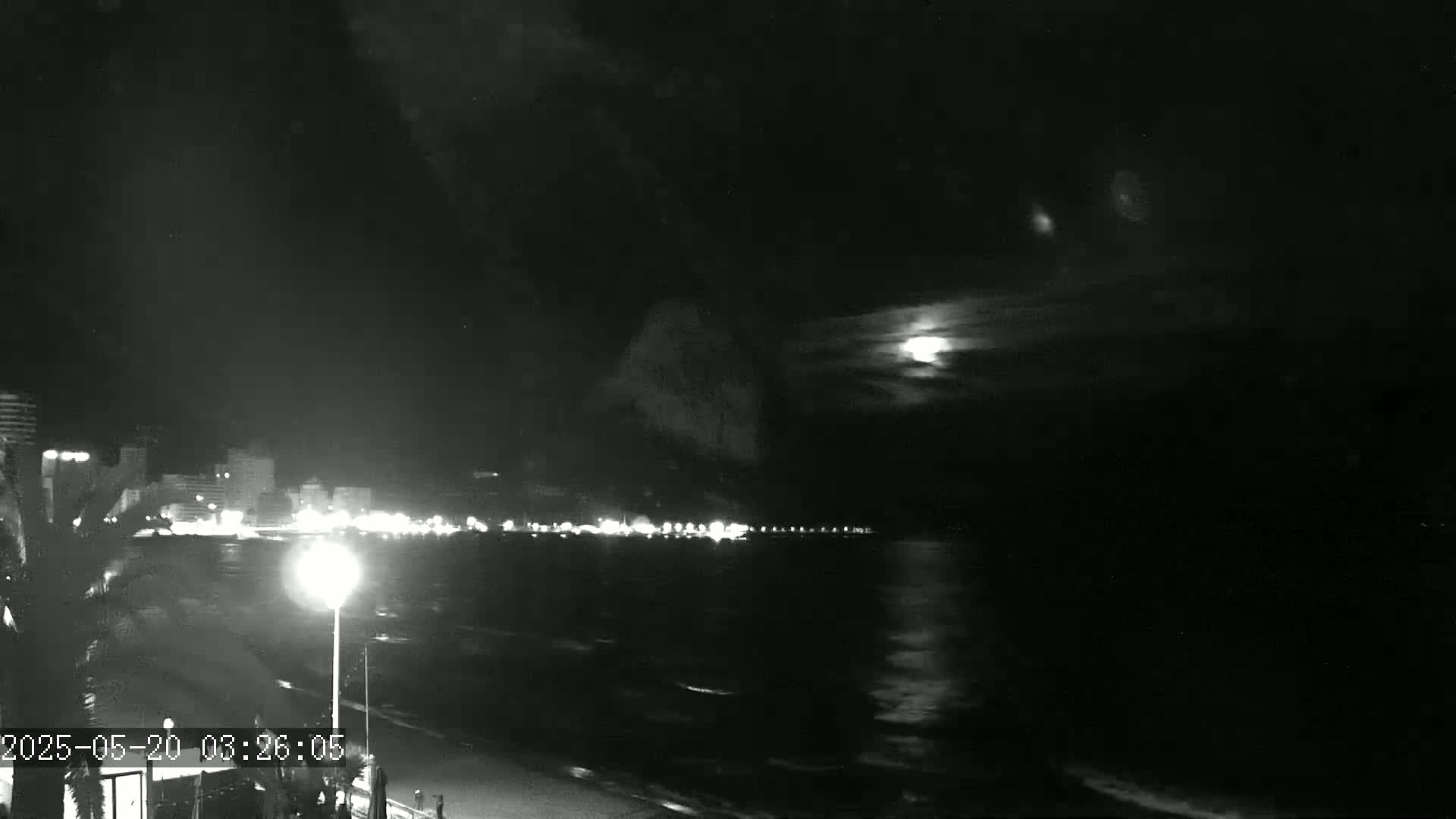 A nighttime, moonlit view of a city's illuminated beachfront and ocean, with a partly cloudy sky.