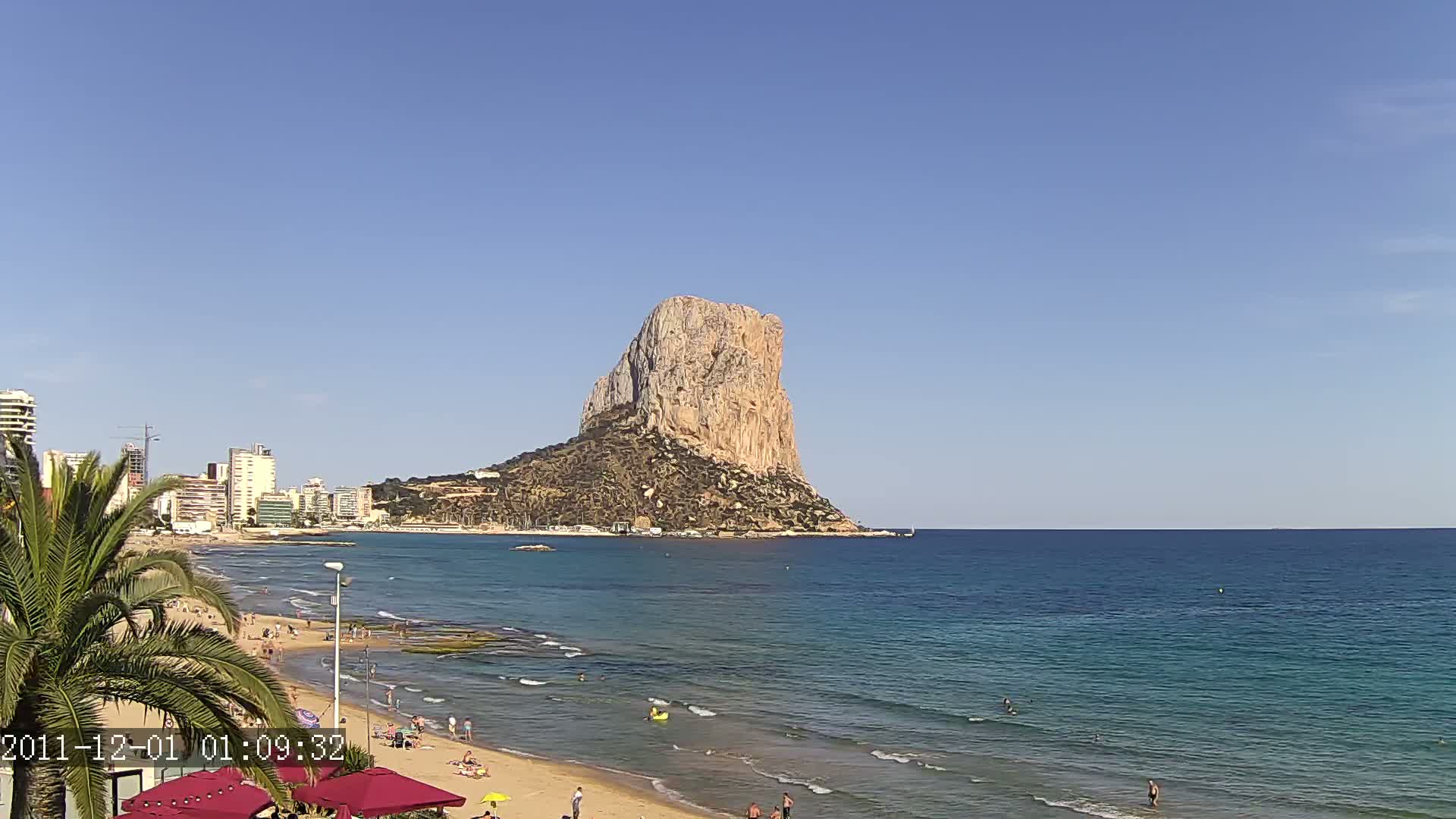 A large, tan rock formation rises from the sea, visible from a sandy beach with people swimming and sunbathing, under a clear blue sky.