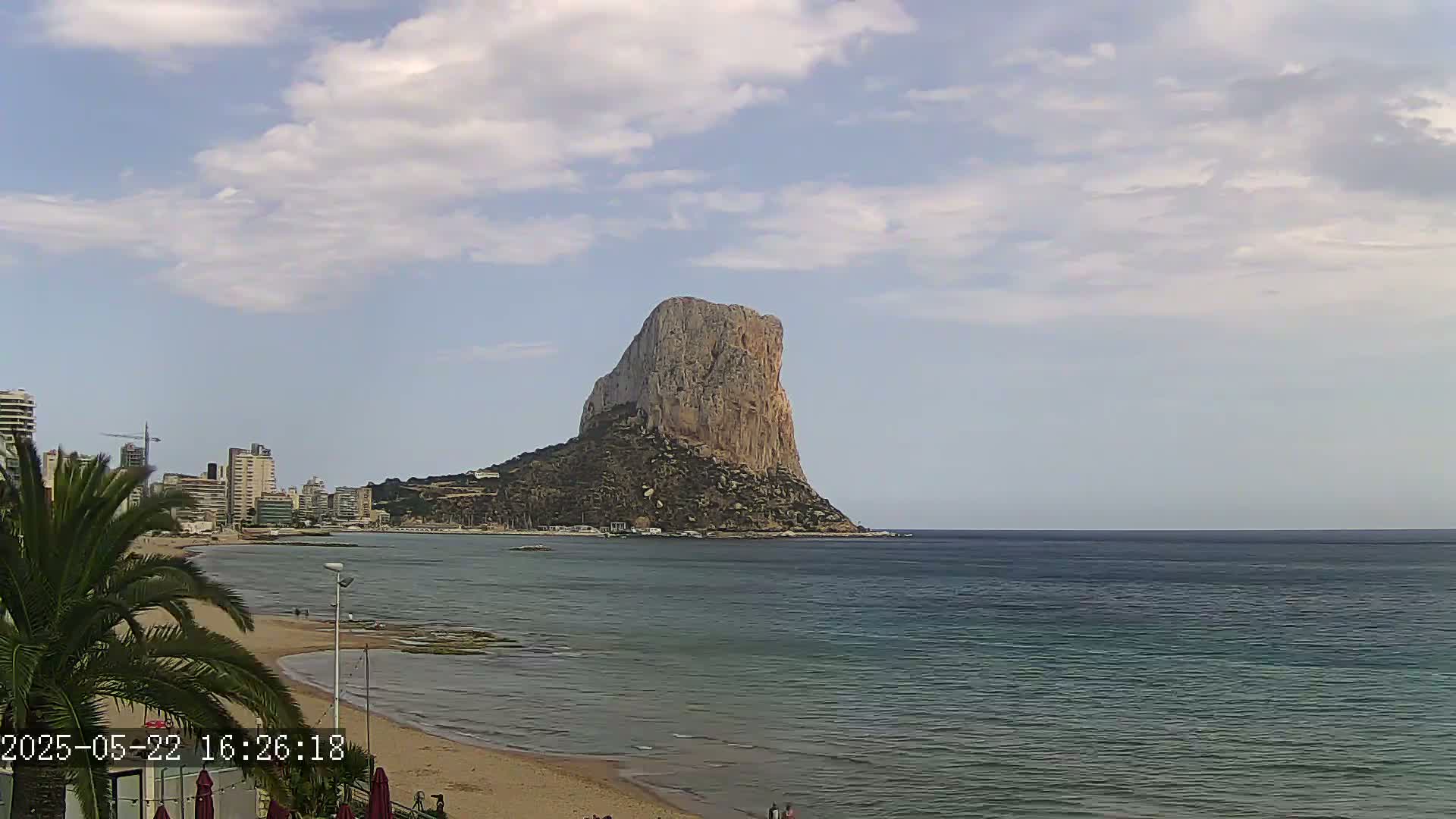 A large, rocky, tan-colored mountain rises from the sea near a sandy beach with a city skyline in the background under a partly cloudy sky.