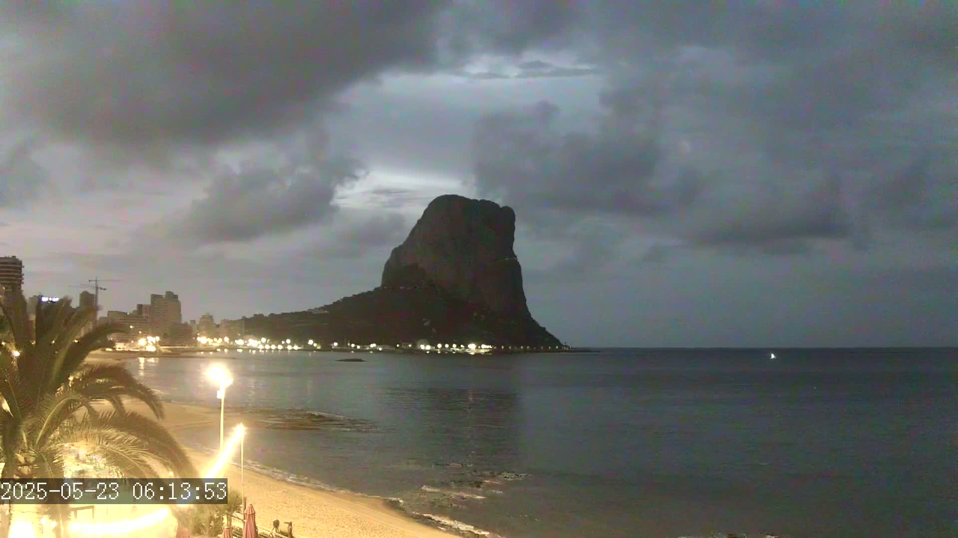 A large, dark rock formation rises from the water at dusk under a cloudy sky, with a dimly lit beach and city skyline visible in the foreground.