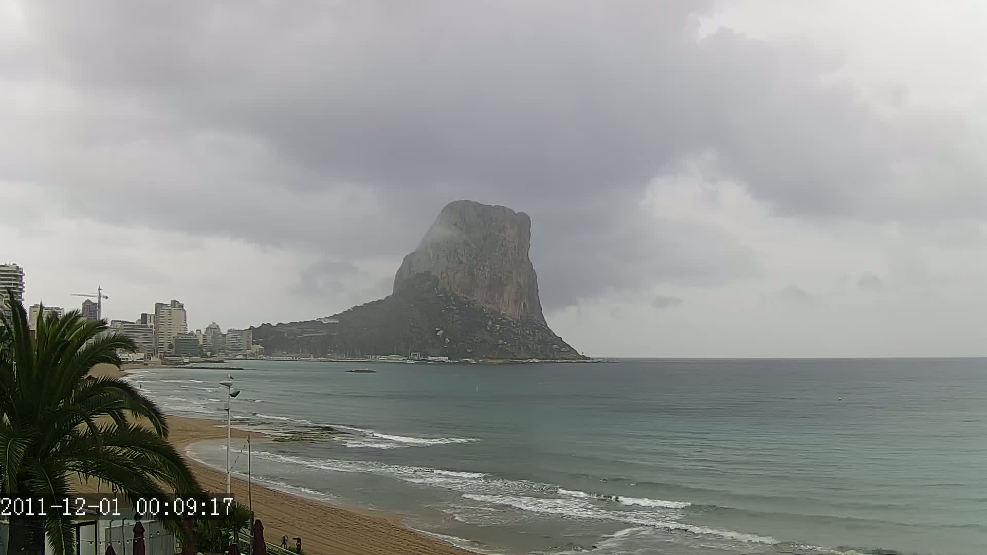 A large, imposing rock formation rises from the sea under a cloudy, overcast sky, with a beach and buildings visible in the foreground.
