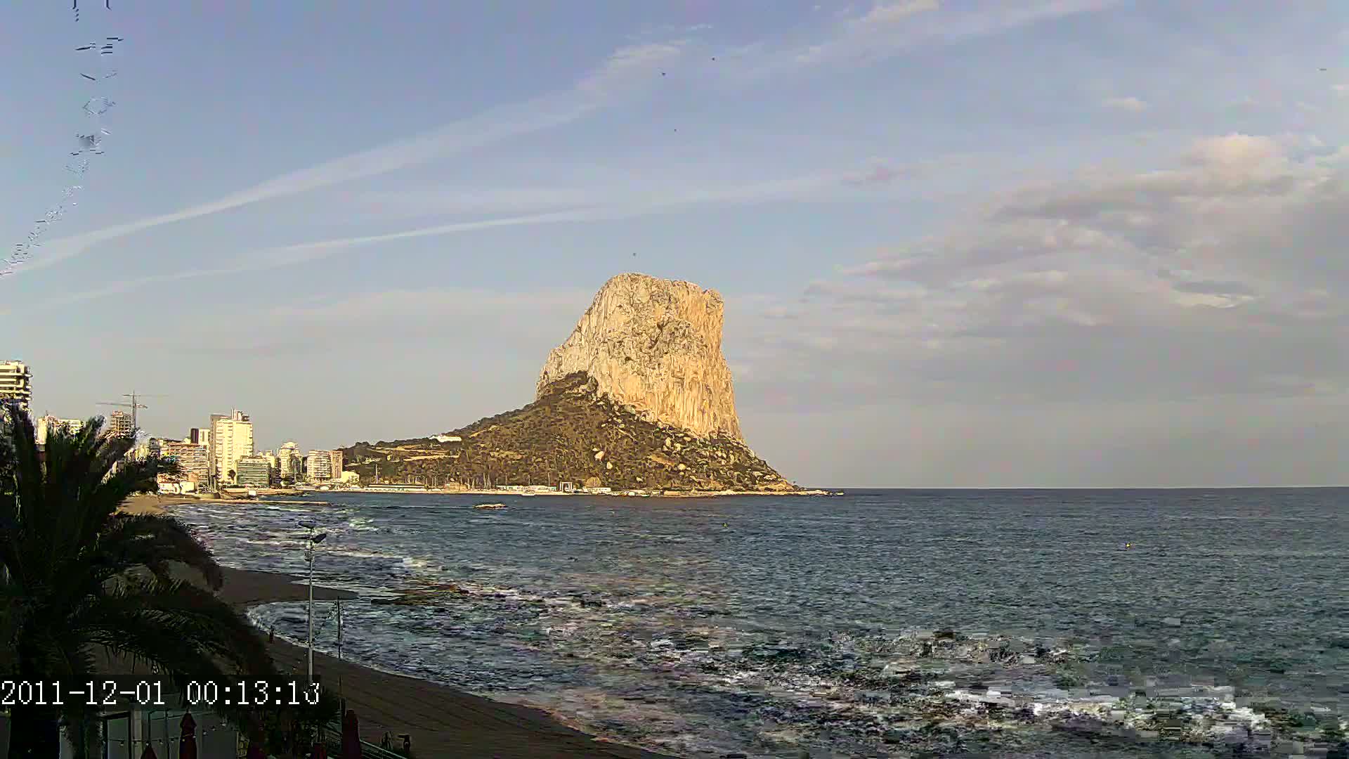 A large, light-colored rock formation rises from the sea under a partly cloudy sky, with a city skyline and beach visible in the foreground.