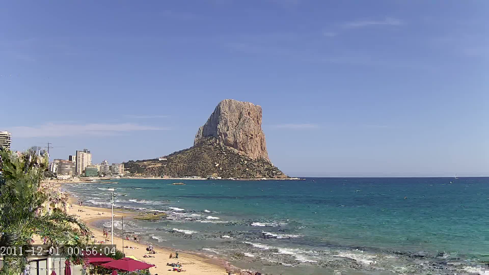A large, rocky outcrop rises from the sea near a sandy beach with people sunbathing under a clear blue sky.