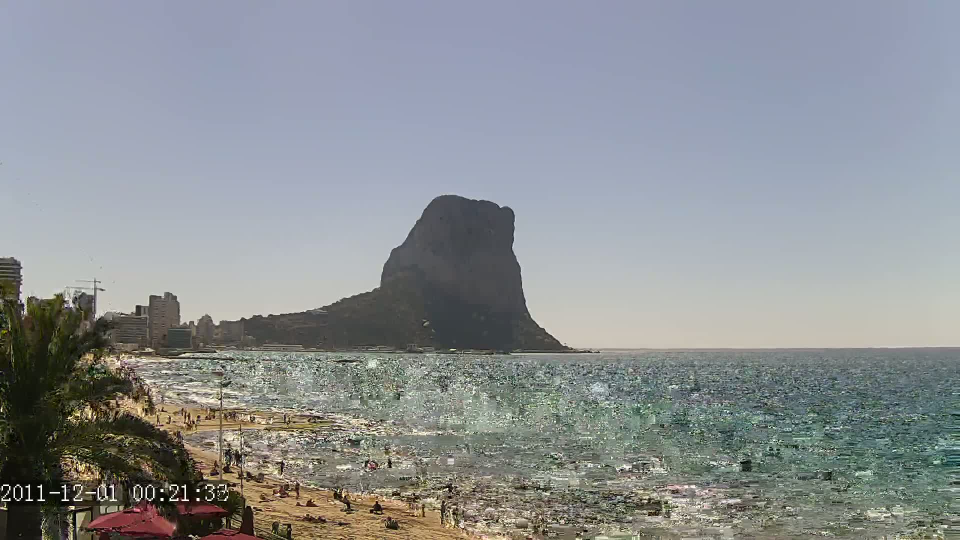 A large, dark rock formation rises from the sea near a sandy beach with people sunbathing and swimming under a clear, sunny sky.
