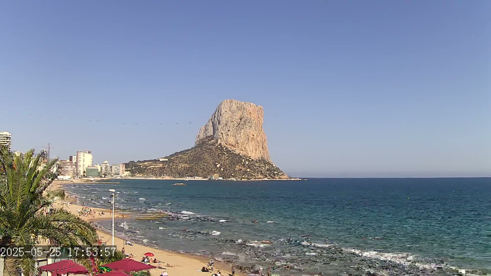 A large, tan rock formation rises from the sea near a sandy beach with people sunbathing and swimming under a clear blue sky.