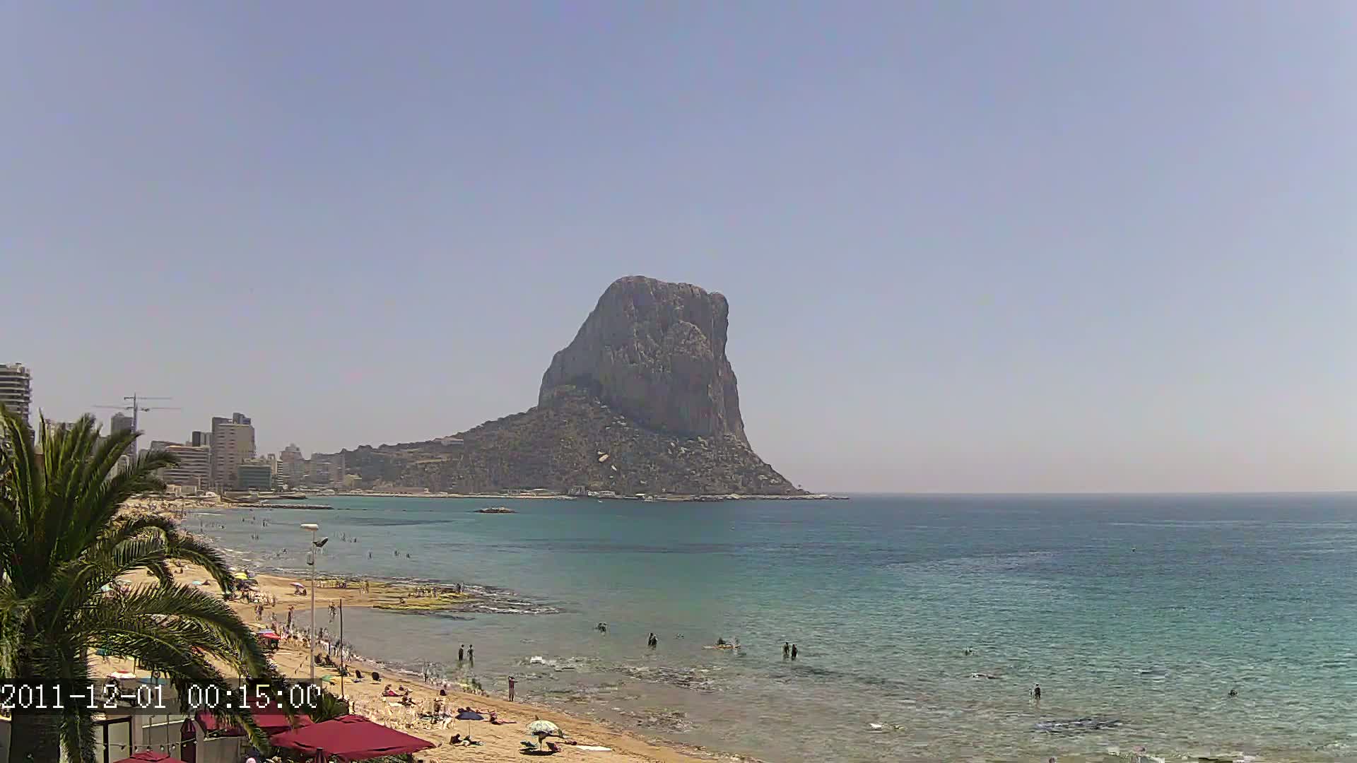 A sandy beach with swimmers in the ocean, under a clear sky, features a large, prominent rock formation in the distance.