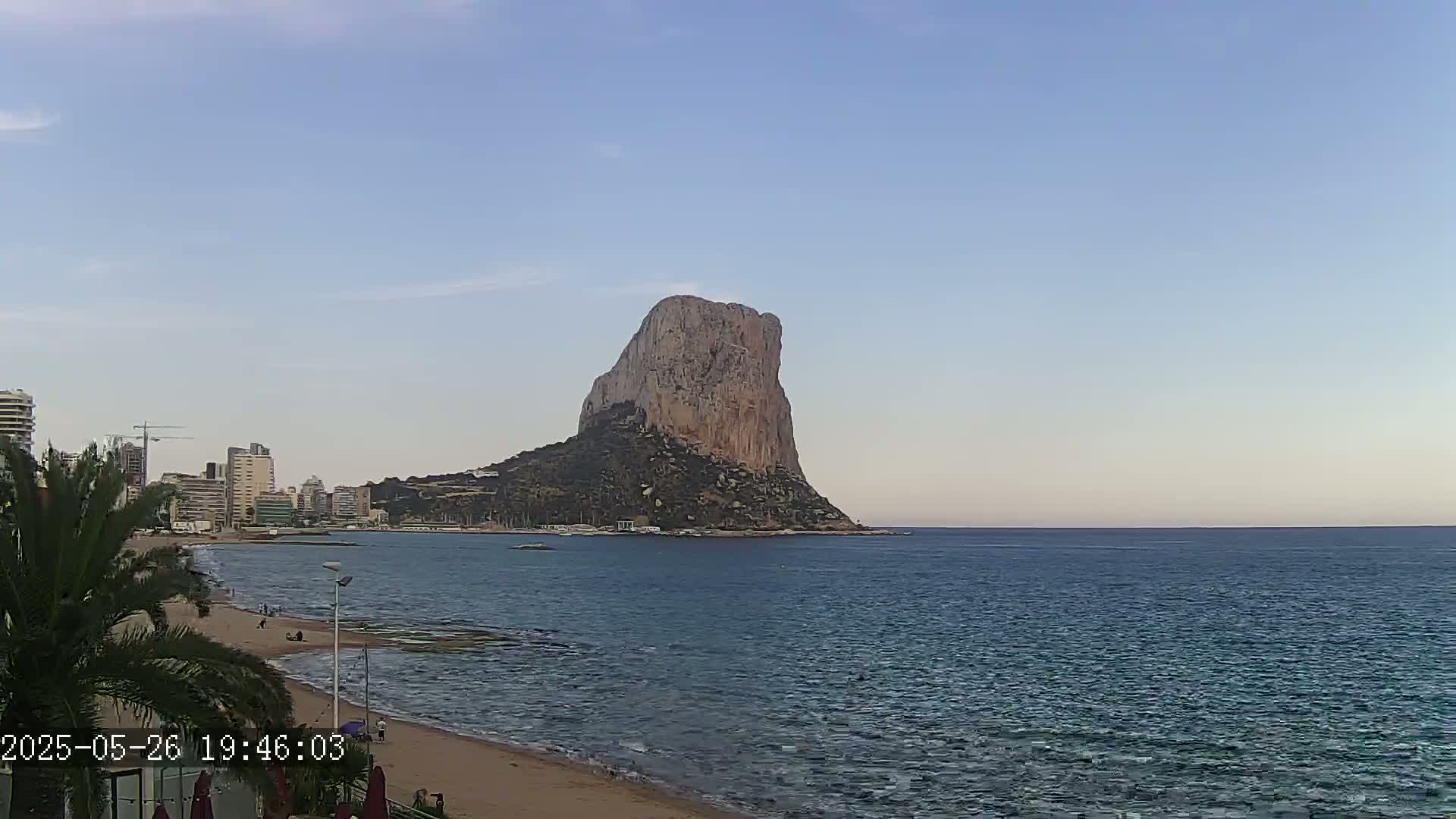 A large, rocky island sits in a calm sea under a clear, light-blue sky, with a beach and a city skyline visible in the foreground.