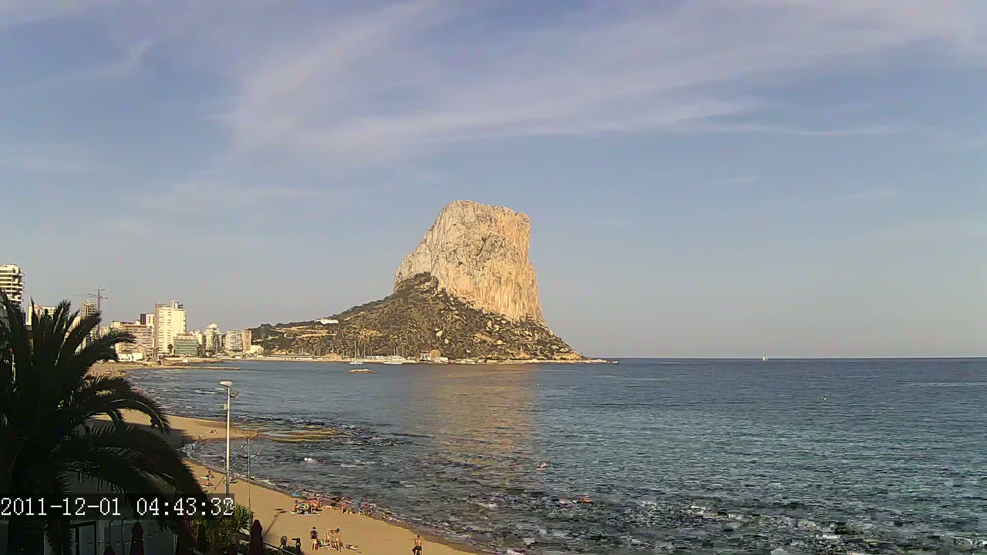 A large, light-brown rock formation rises from the sea under a mostly clear, light-blue sky, with a sandy beach and buildings visible in the foreground.