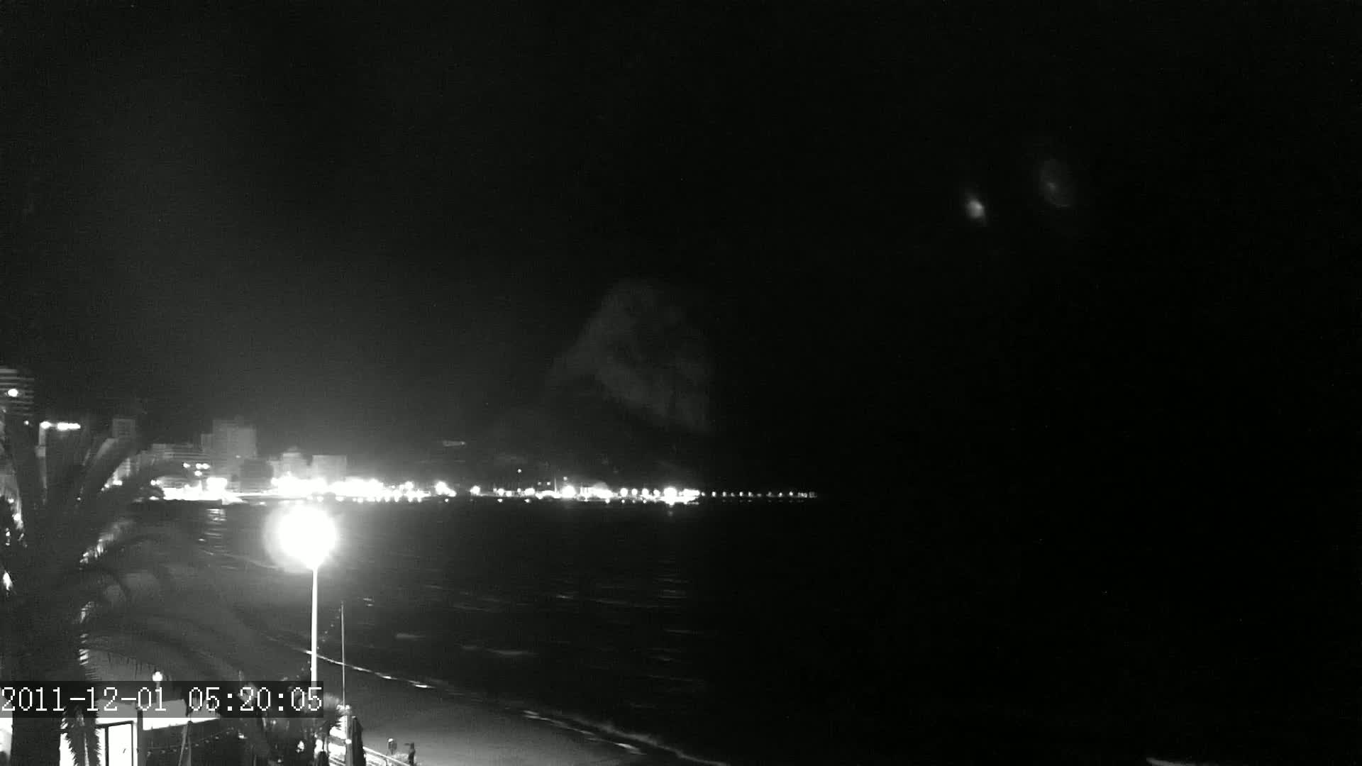 A nighttime view of a coastline with city lights illuminating the water and a dark mountain in the background under a clear, dark sky.