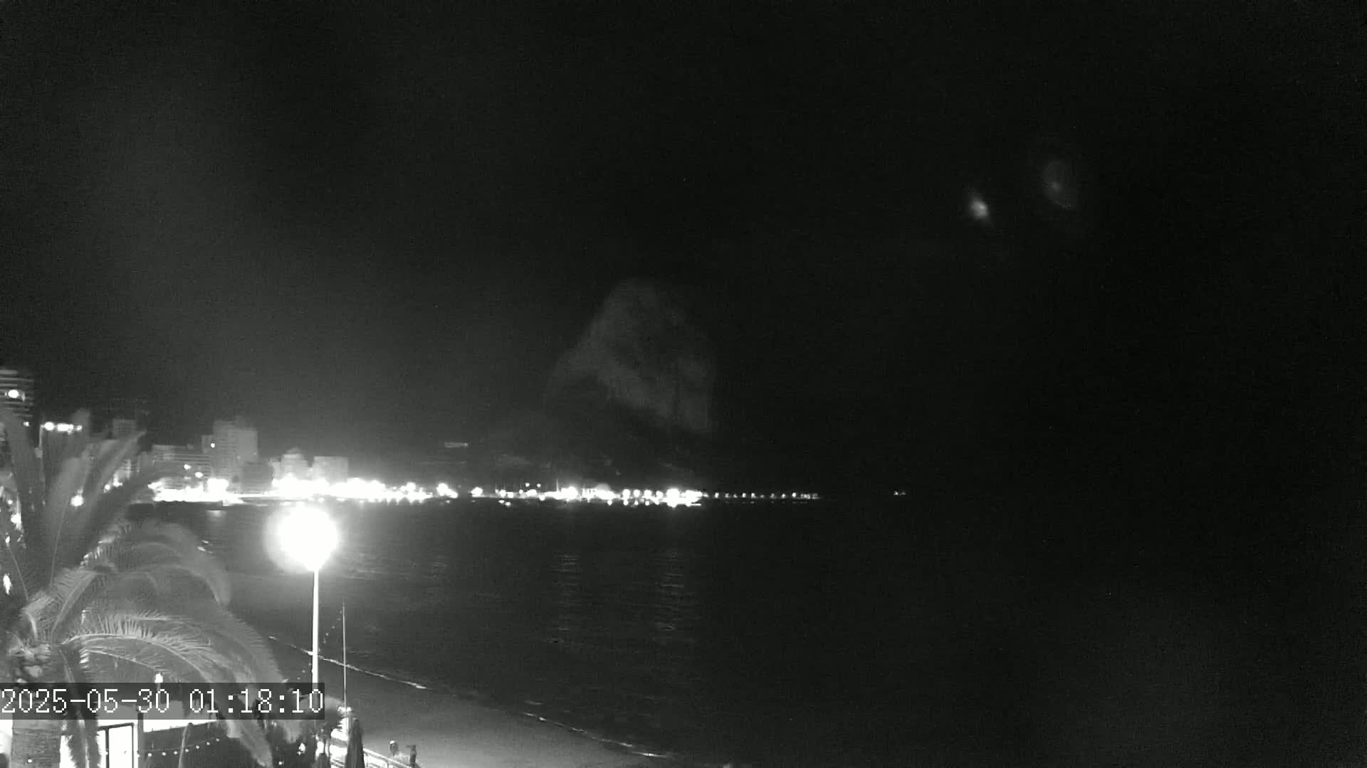 A nighttime coastal scene shows a dark, rocky outcrop across a calm body of water from a city's illuminated shoreline under a clear, dark sky.