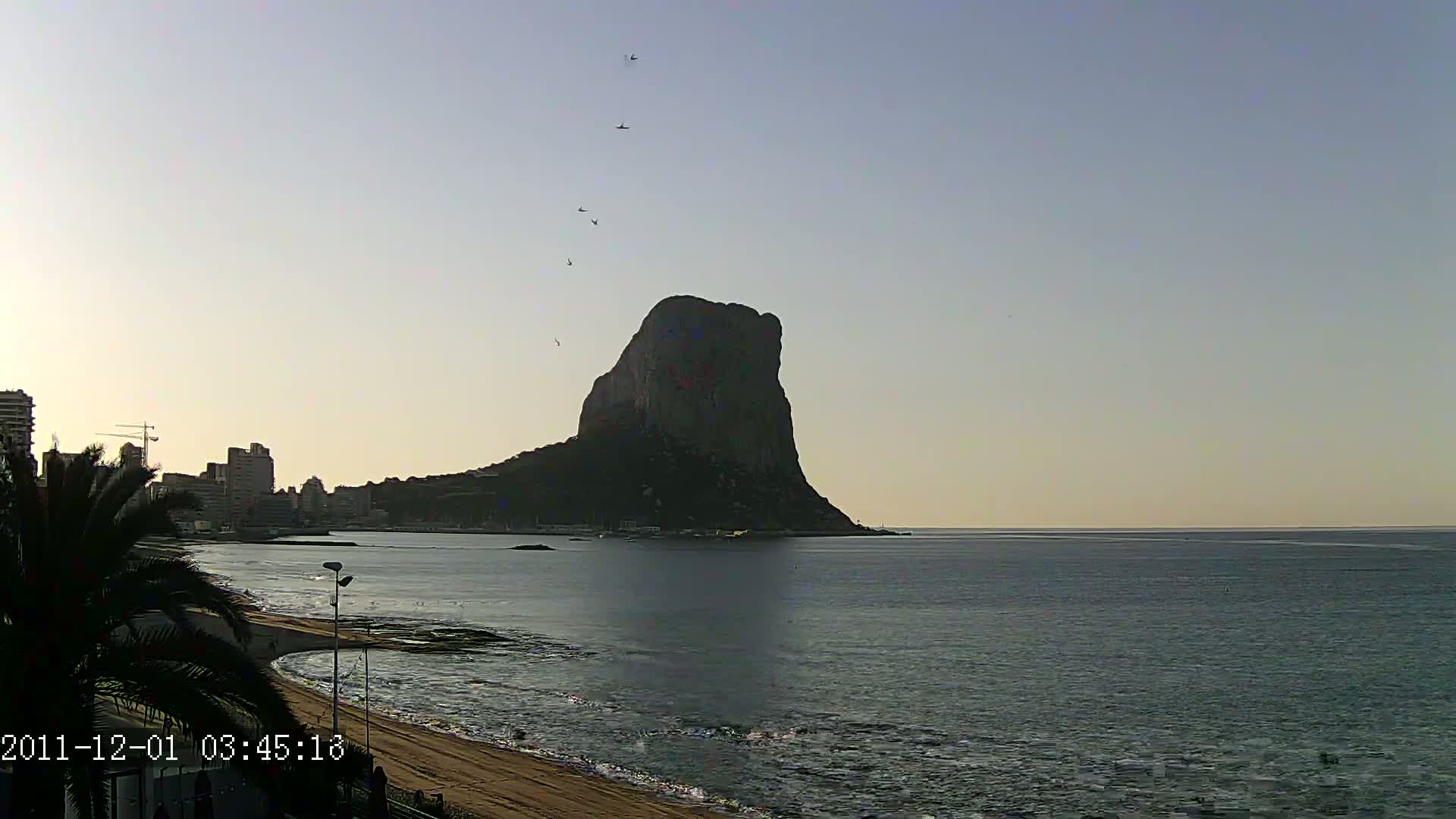 A large, dark rock formation rises from the calm sea under a pale, clear sky, with a city skyline visible in the background and a sandy beach in the foreground.