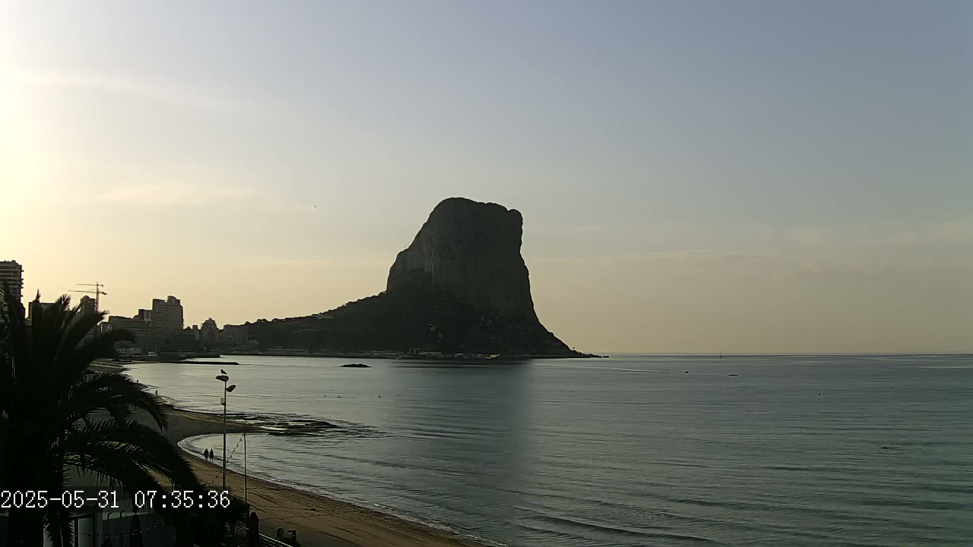 A large, dark rock formation rises from the calm, gray-blue sea near a sandy beach and a cityscape under a clear, pale sky.