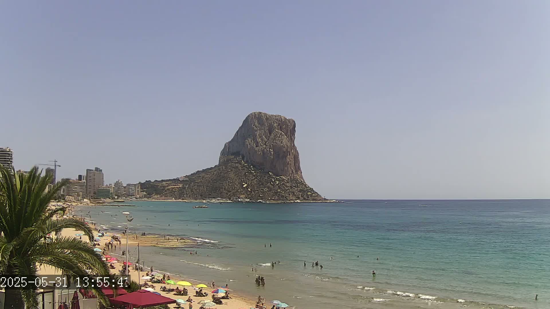 A sandy beach with people sunbathing and swimming,  backed by buildings, and overlooking a large, rocky,  island in a calm, sunny ocean.