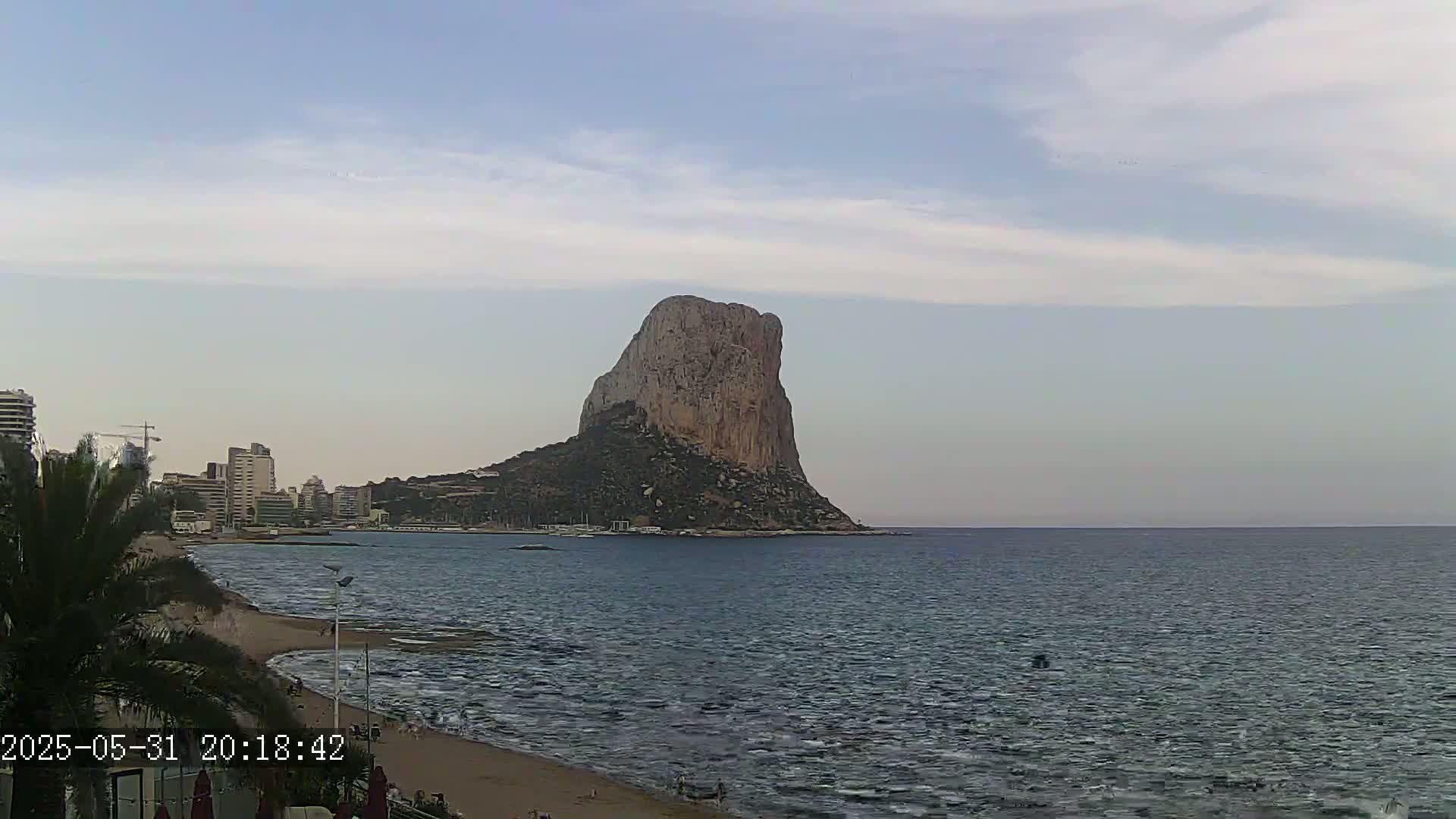 A large, rocky, and isolated hill rises from the sea under a mostly clear, light-blue sky, with a sandy beach and buildings visible in the foreground.