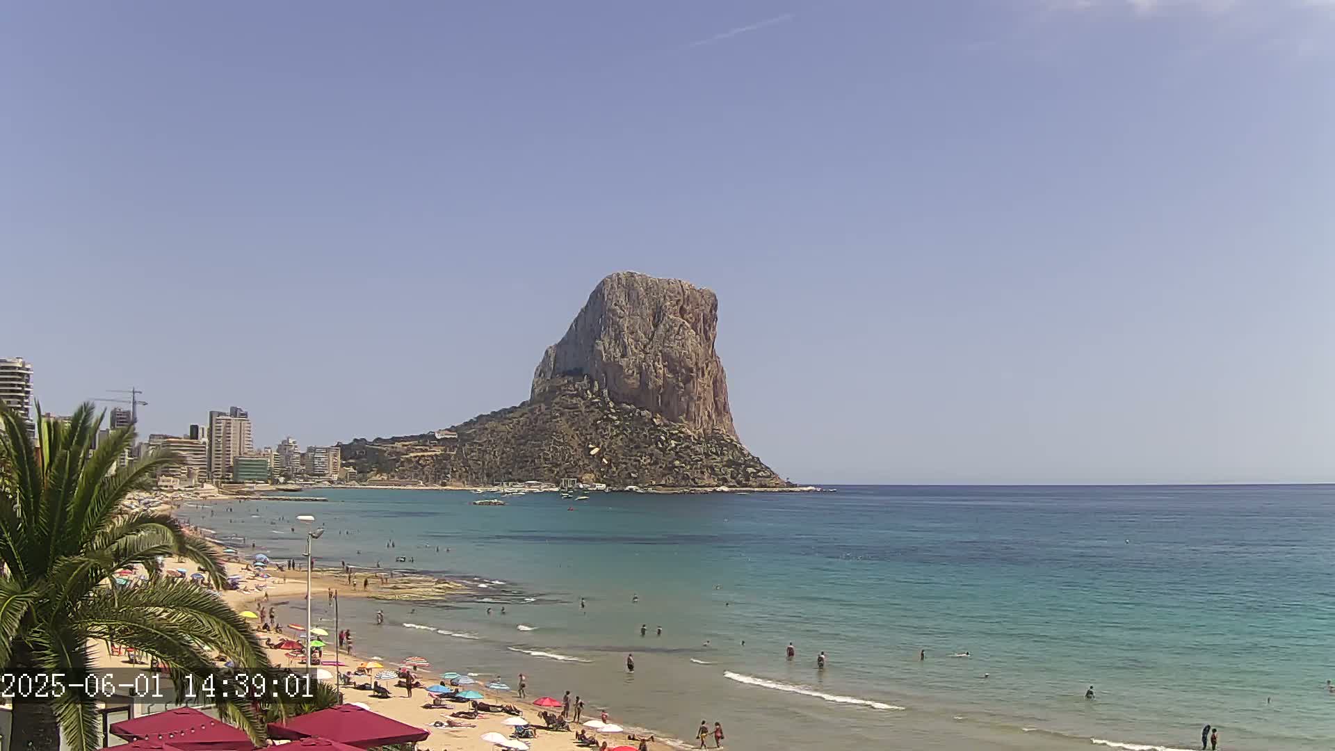 A large rock formation rises from the sea on a sunny day, overlooking a beach with people swimming and sunbathing.