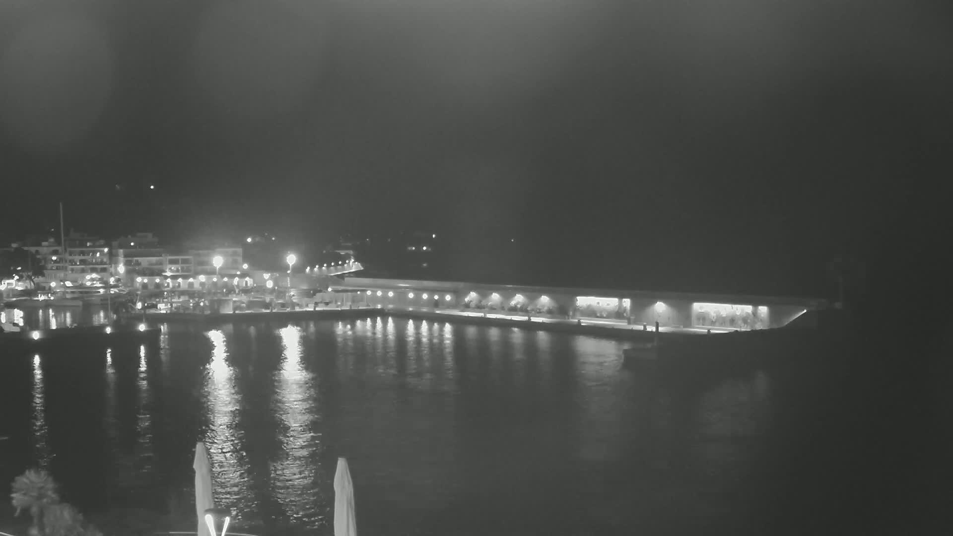 A nighttime view of a harbor with illuminated buildings and boats, reflecting lights on calm water under a dark sky.