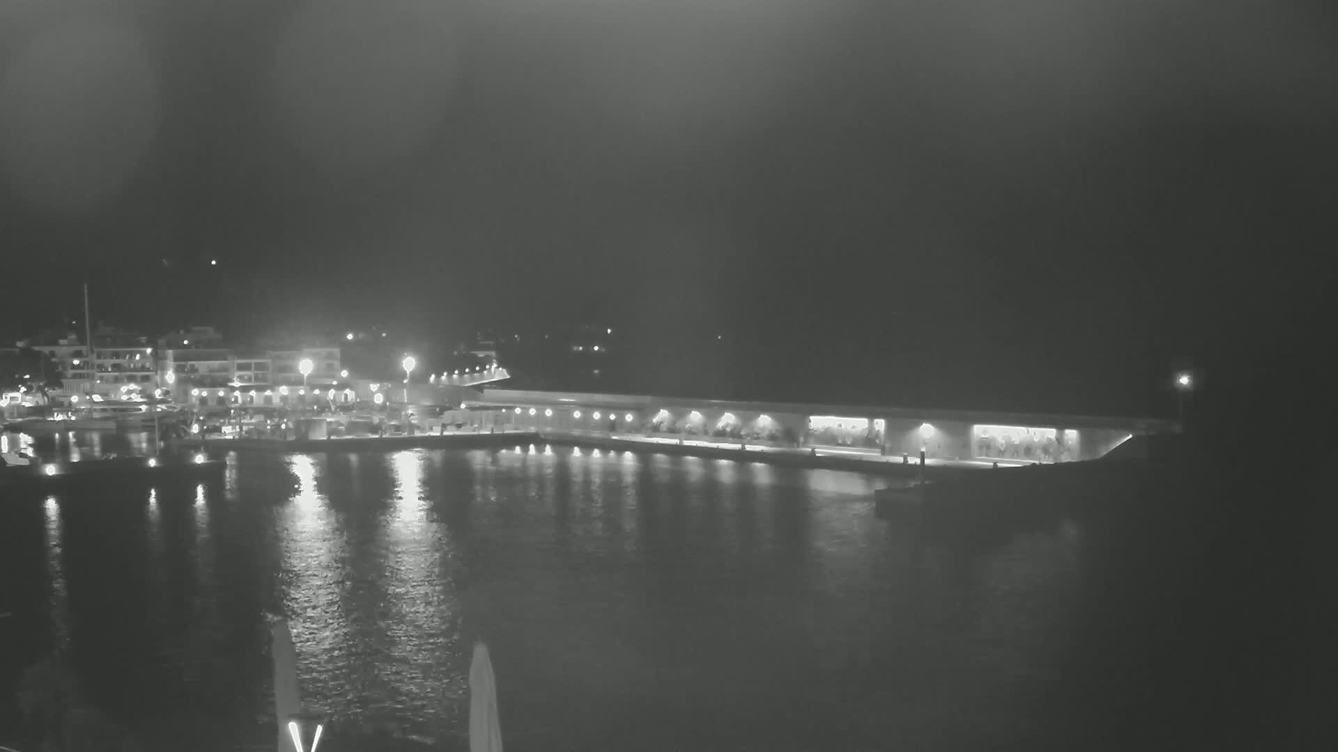 A nighttime view of a harbor, illuminated by lights reflecting on calm water, shows buildings along the shoreline.