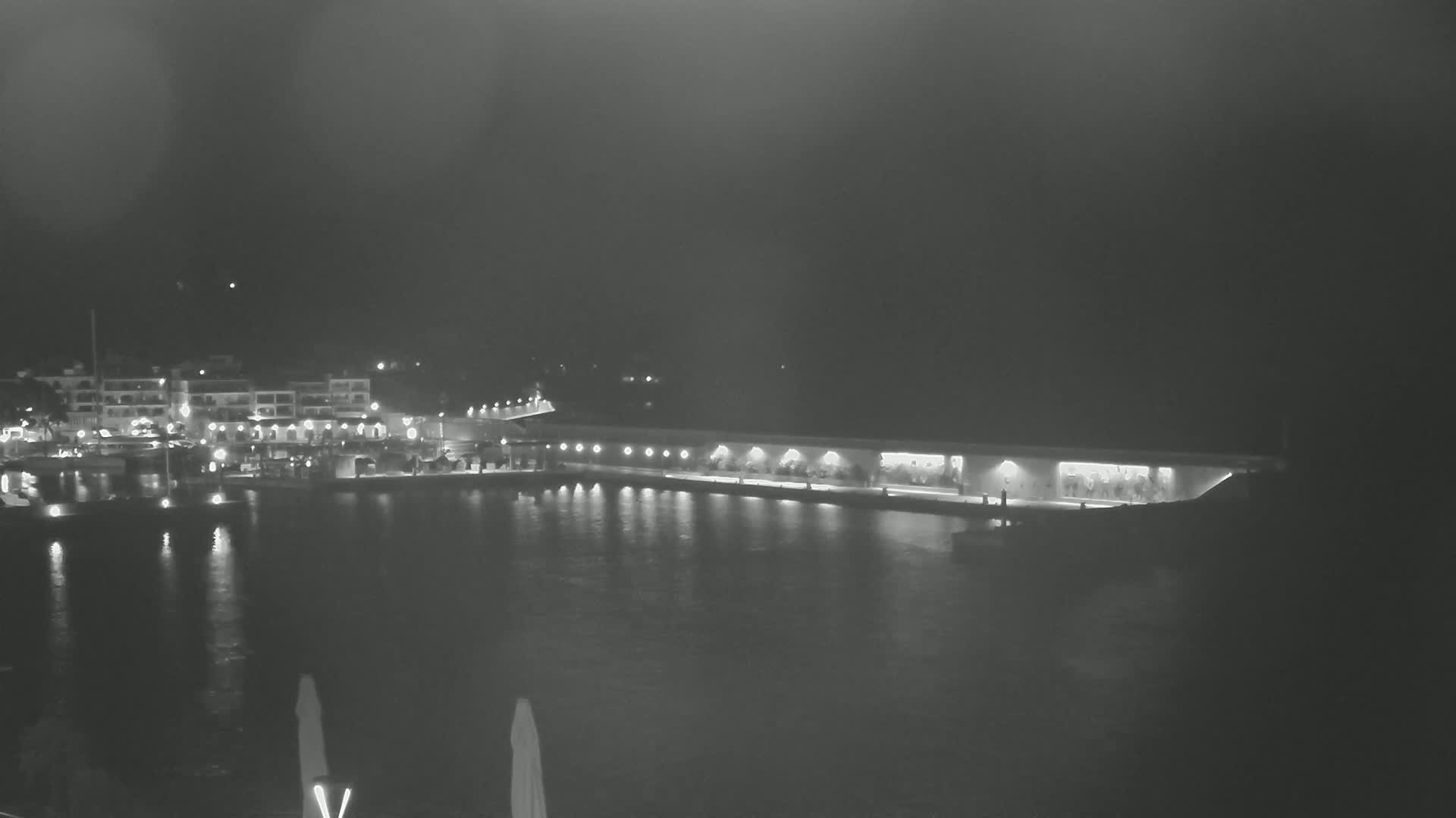 A nighttime view of a harbor with a long, illuminated building along the waterfront, under a hazy sky.