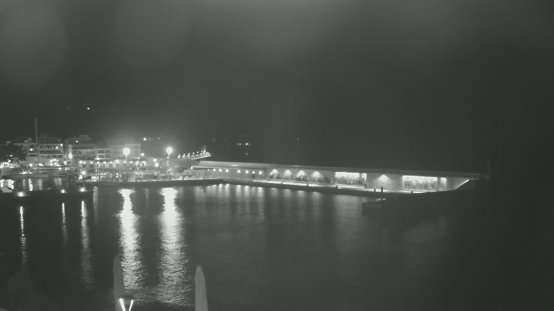 A nighttime harbor scene shows a long, low building with lights along a waterfront, reflecting in calm water, with boats and buildings visible across the water.
