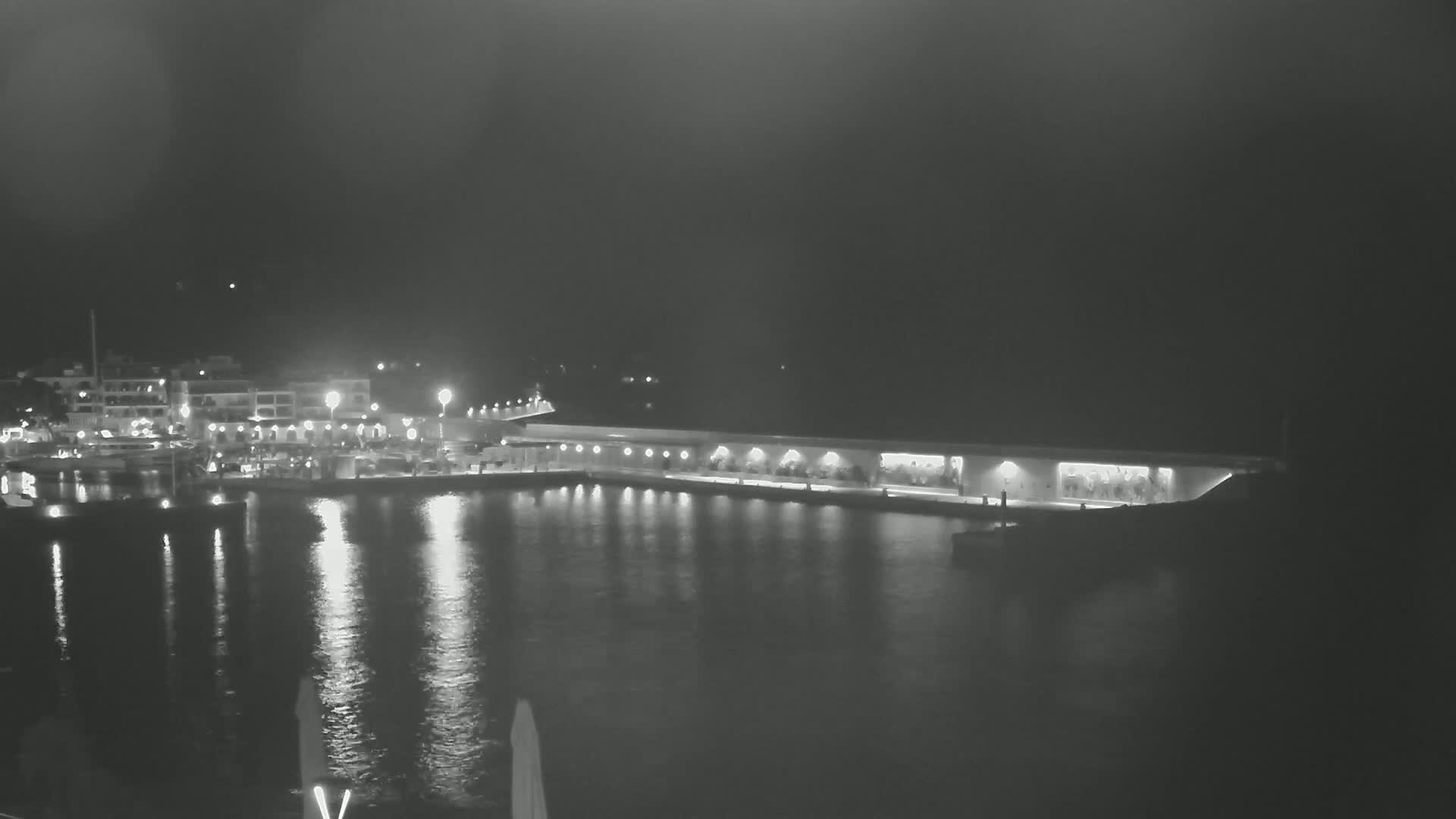 A nighttime view of a harbor with a long, lit structure extending into the water, boats in the marina, and buildings along the shoreline.