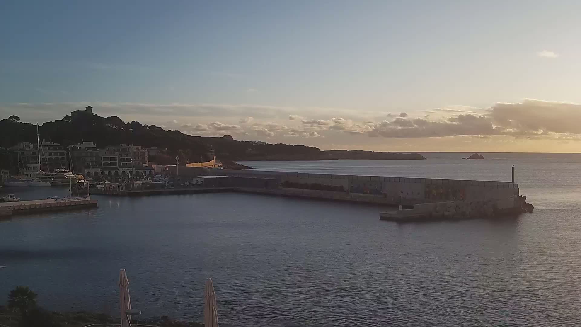 A calm sea at sunset, with a harbor filled with boats nestled next to a hillside town.