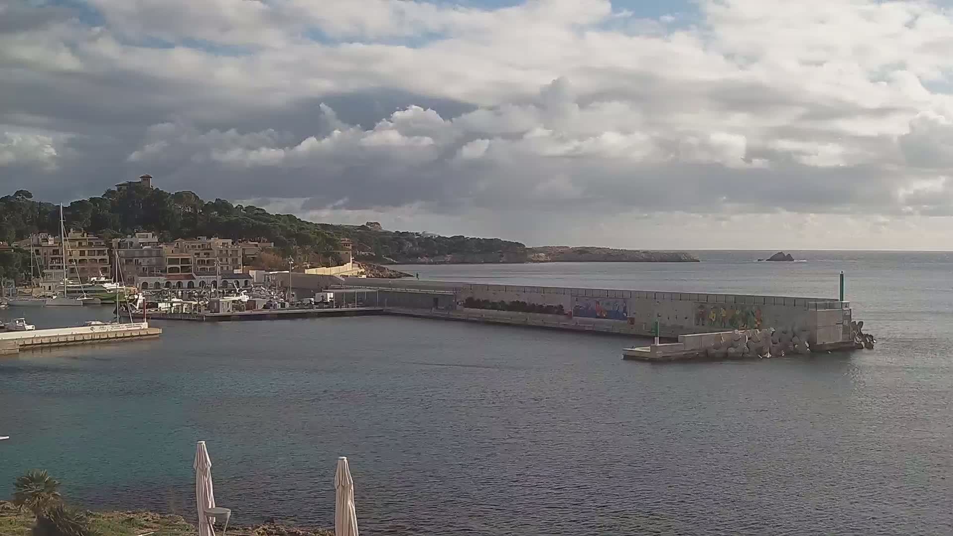 A harbor filled with boats is nestled next to a hilly coastline under a partly cloudy sky.