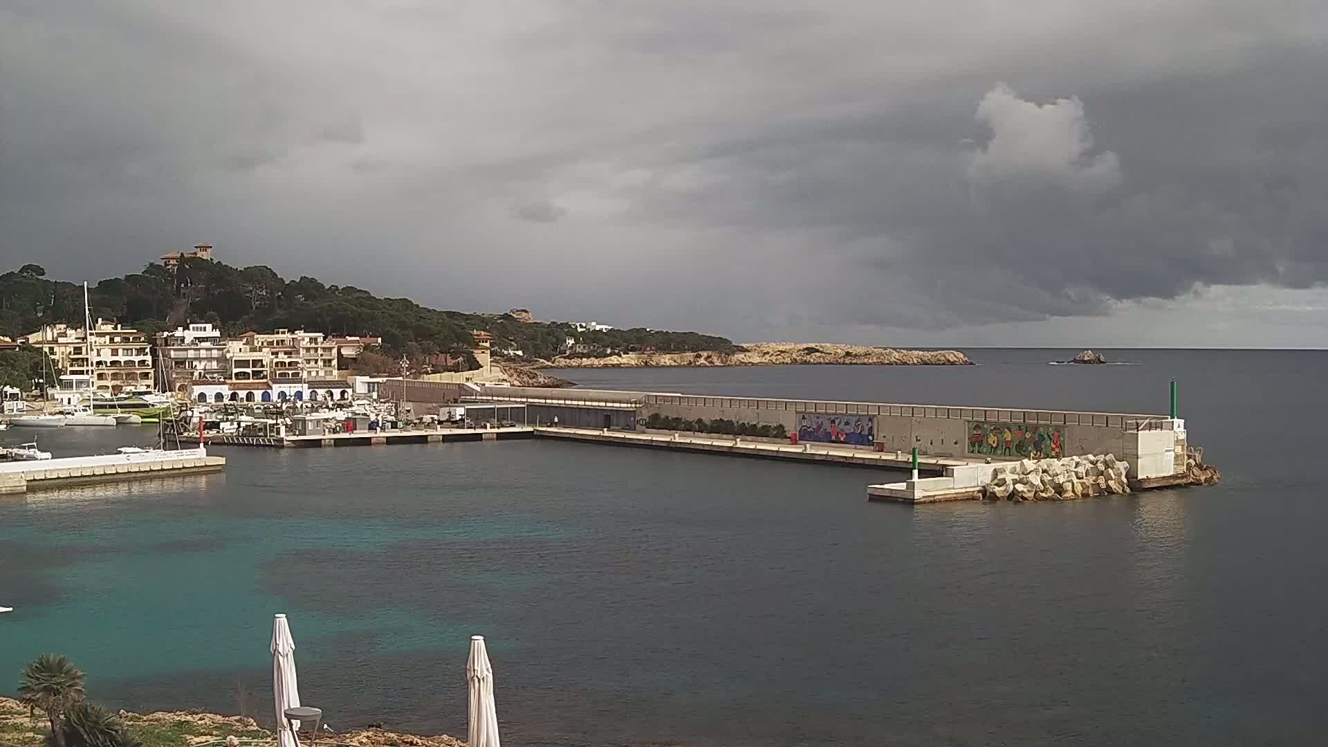 A small harbor with boats and buildings on a hill under a cloudy sky.