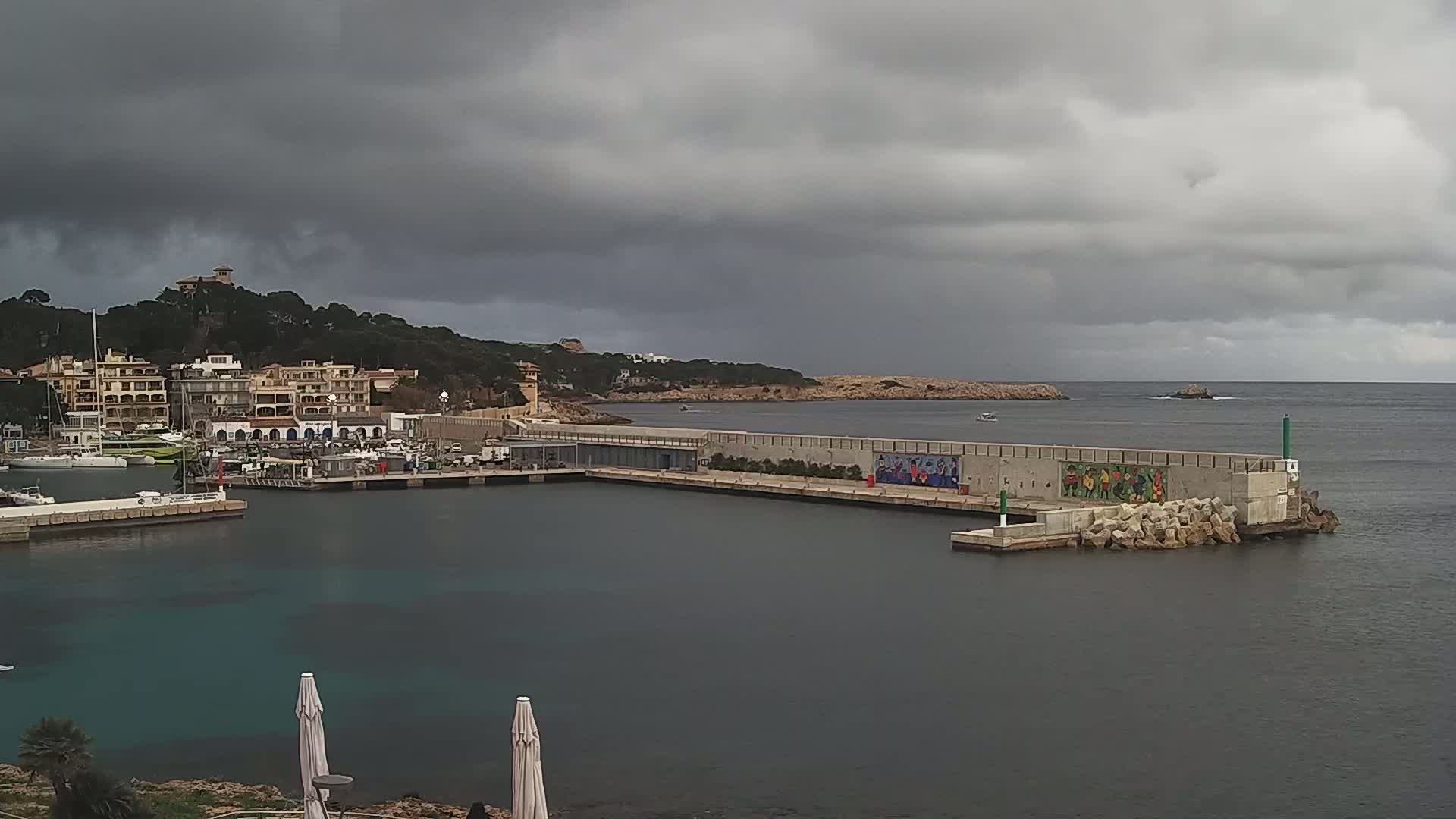 A harbor filled with boats sits under a gray, overcast sky next to a coastal town nestled against a hillside.