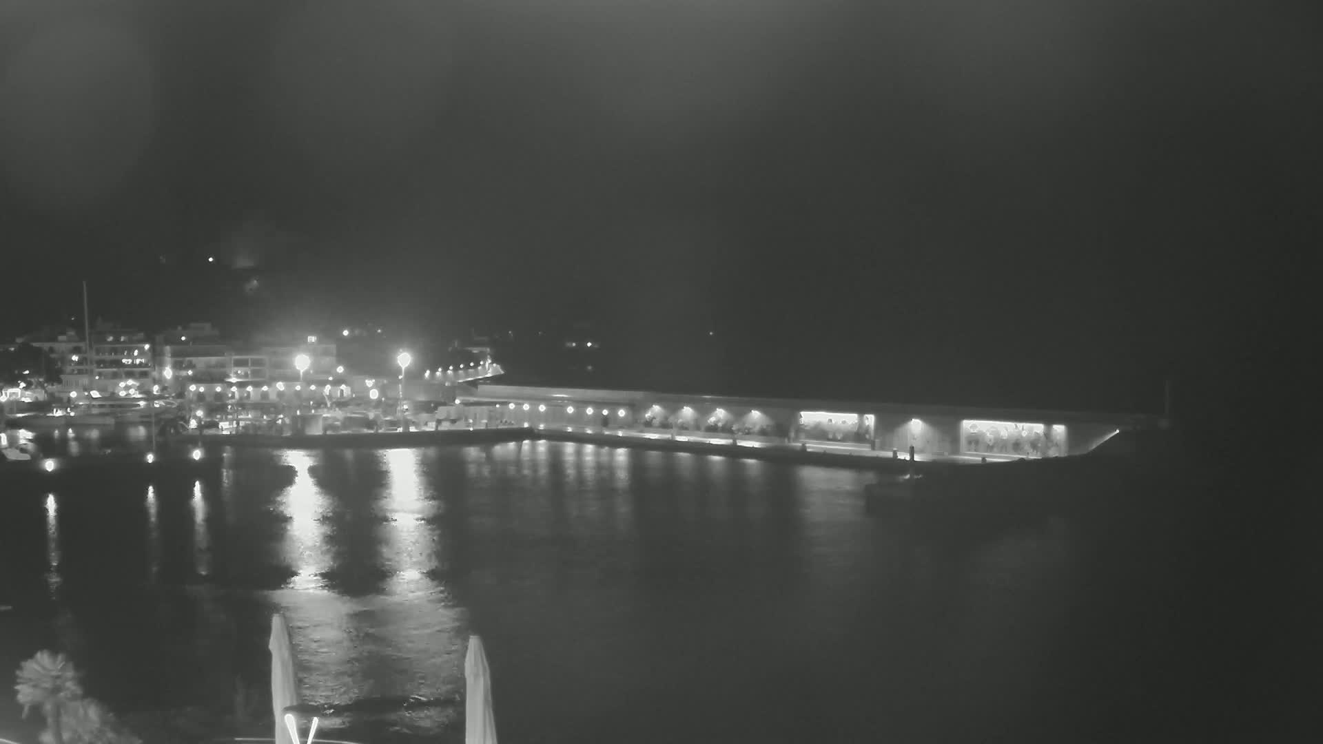 A nighttime view of a harbor with lights reflecting on calm water, showing buildings and boats along the shoreline.