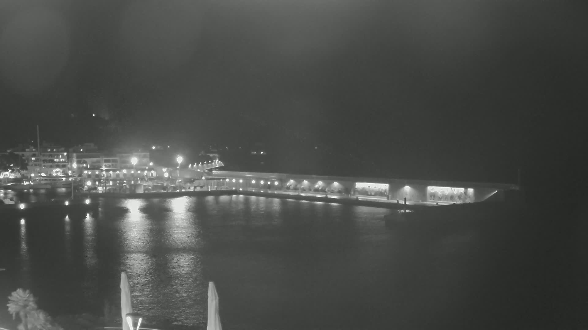 A nighttime view of a harbor with boats moored at a waterfront building, illuminated by lights, under a dark sky.