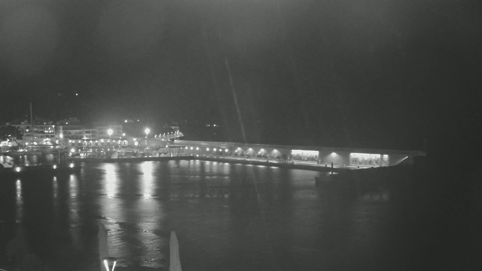 A nighttime, low-light view of a harbor with a long, illuminated waterfront structure and calm water reflecting the lights from buildings across the water.