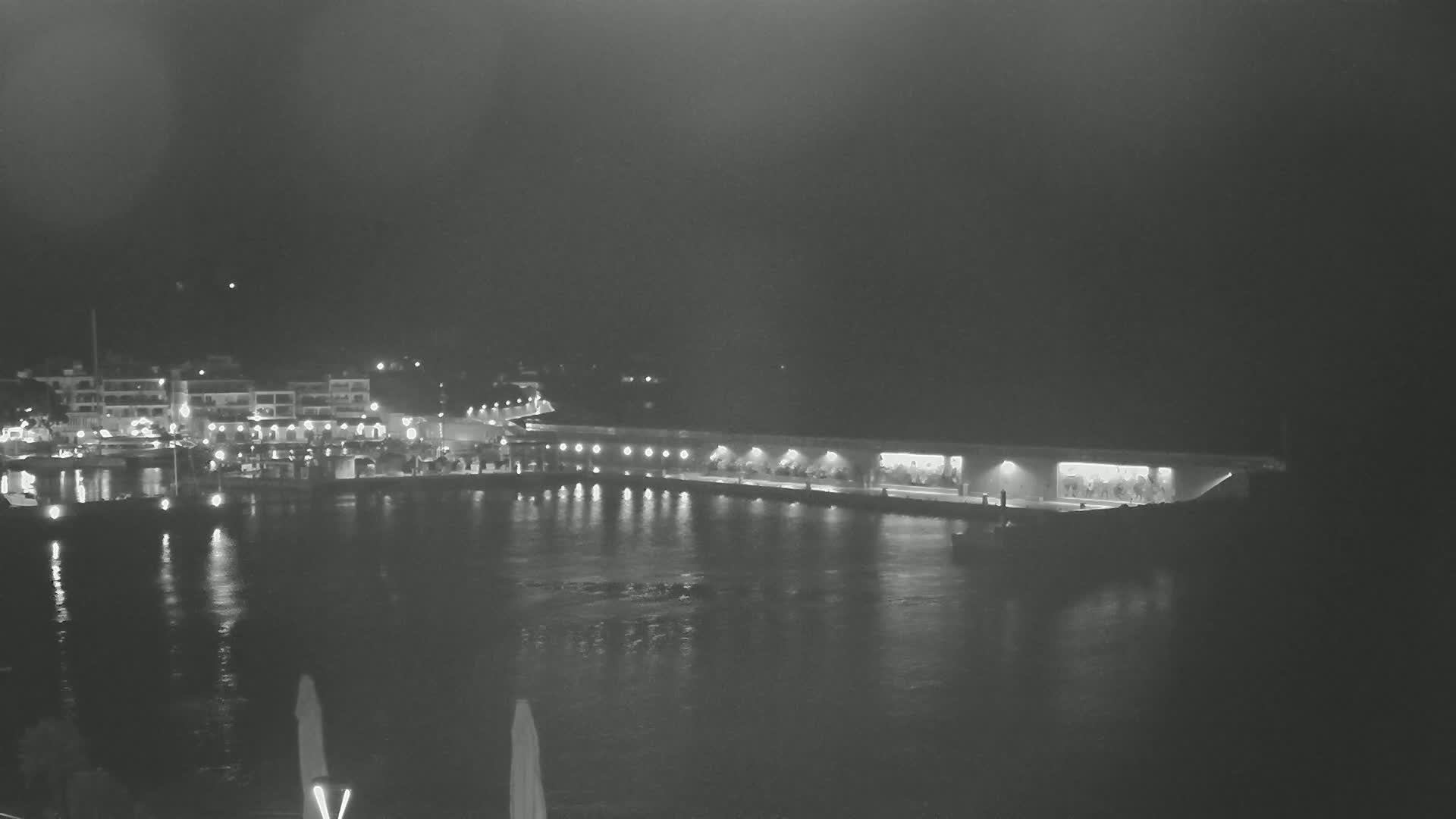 A nighttime view of a harbor with boats and buildings illuminated, under a dark sky.