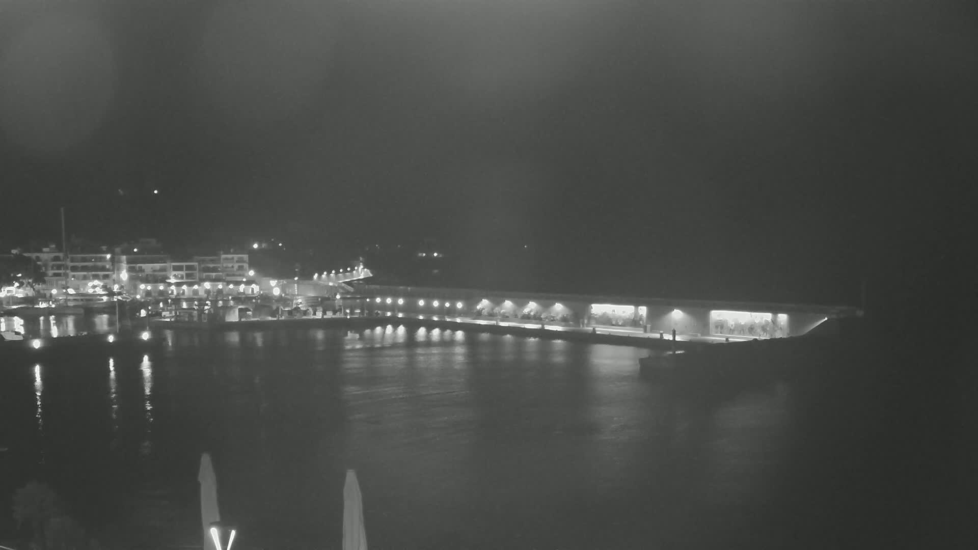 A nighttime view of a harbor with a long pier illuminated, boats in the water, and buildings with lights on the shore.