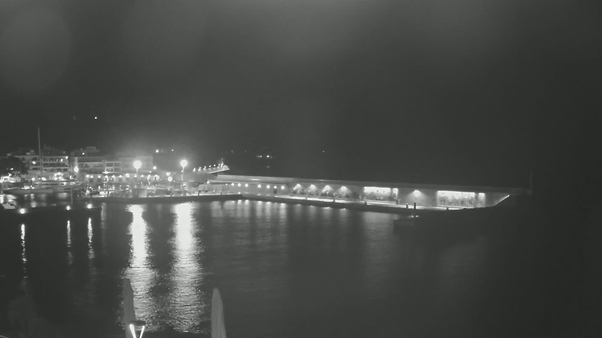 A nighttime harbor scene shows a long, low-lit building along a calm body of water reflecting lights from buildings and boats on the opposite shore.