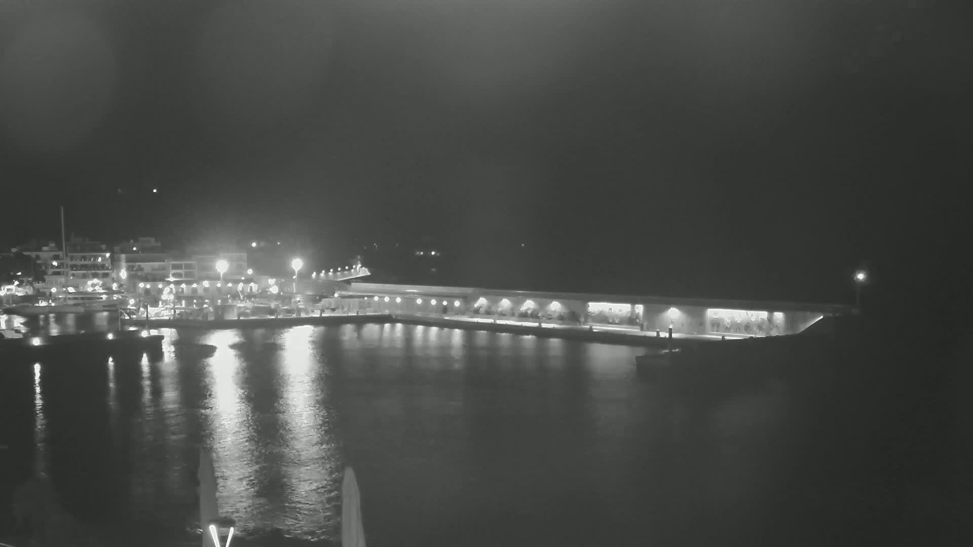 A nighttime view of a harbor with boats docked, buildings lit up along the waterfront, and calm water reflecting the lights.
