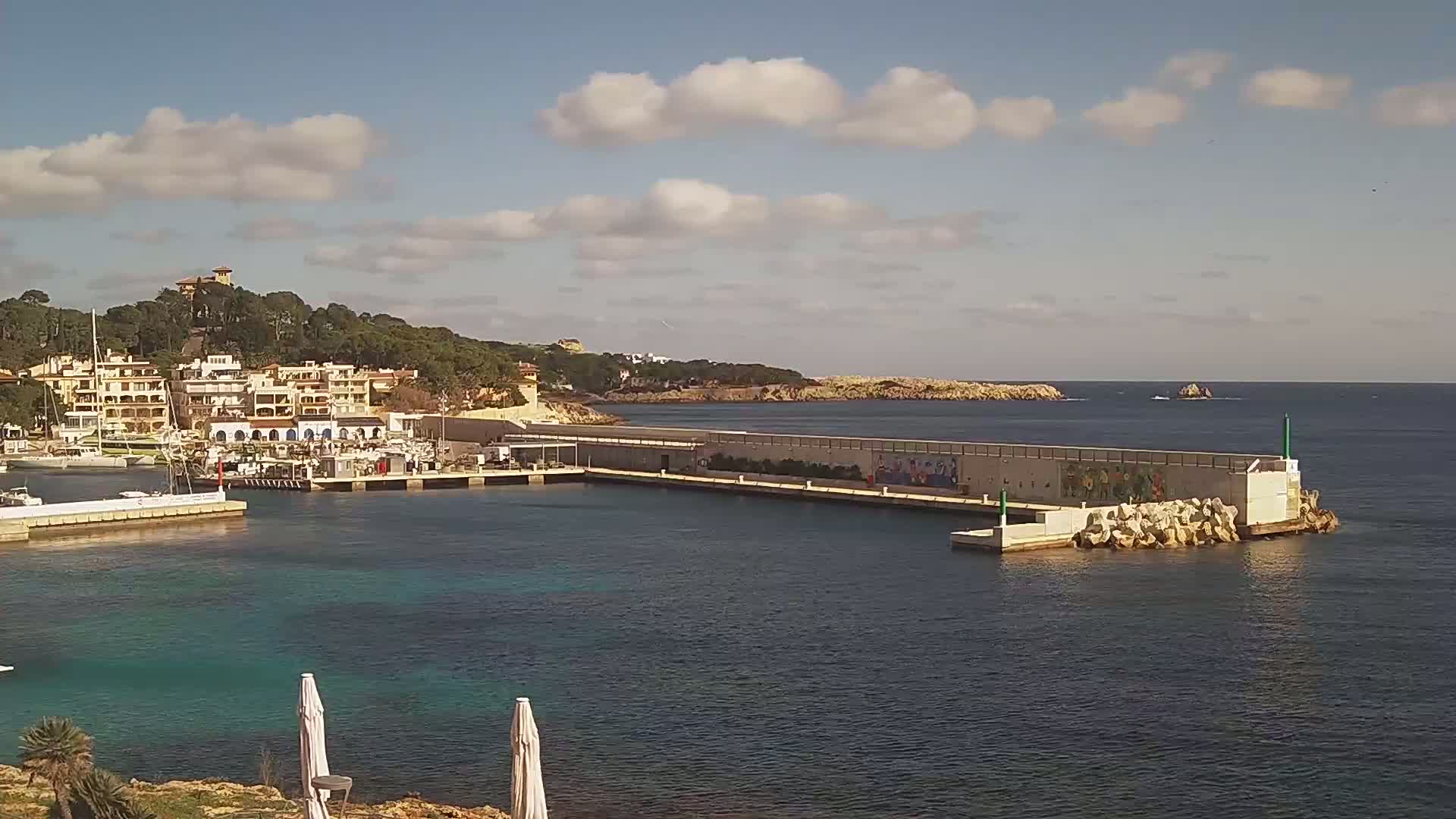 A harbor with a calm sea under a partly cloudy sky, featuring a marina filled with boats and buildings along the shoreline.