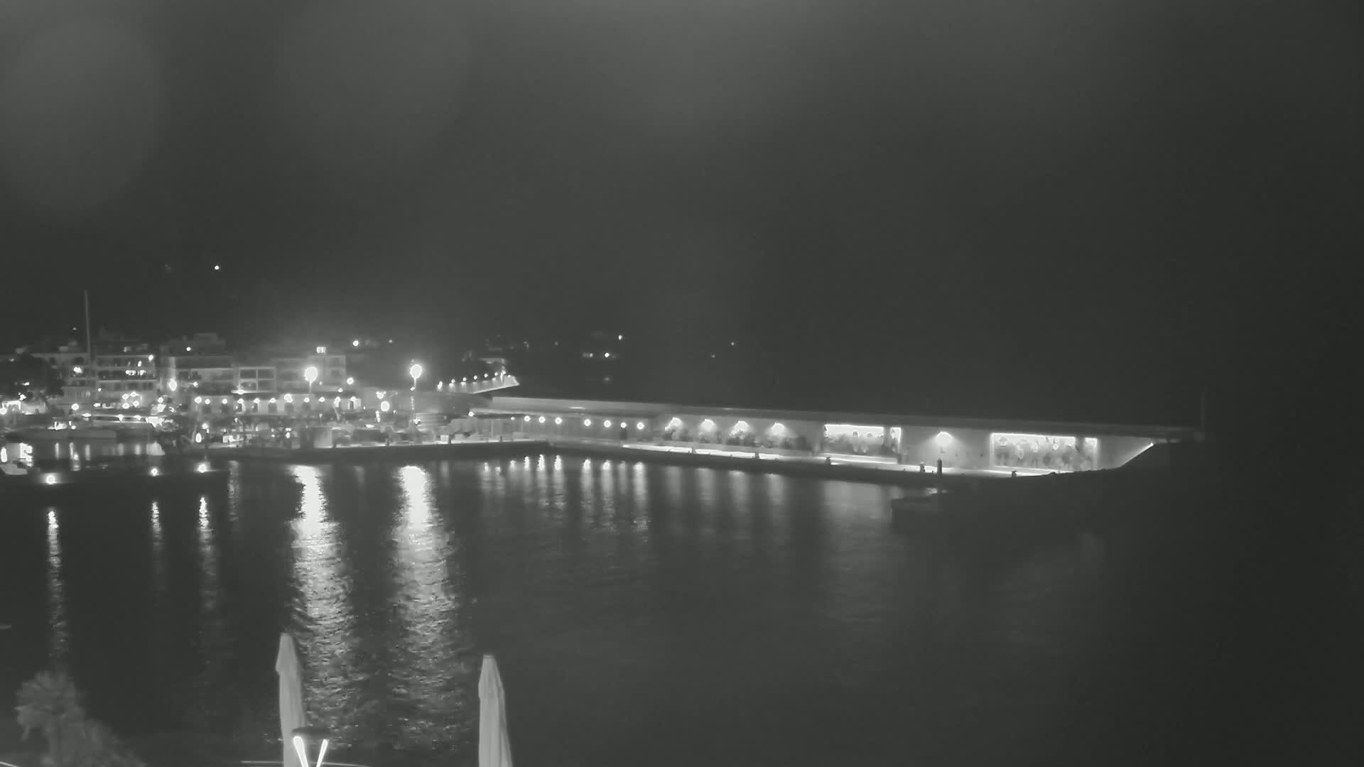 A nighttime view of a harbor, illuminated by lights reflecting on calm water, shows a long building along the waterfront and buildings across the water.