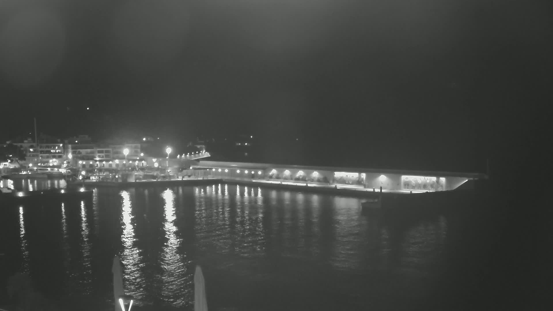 A nighttime view of a harbor with a long, lit waterfront structure, boats, and buildings reflected in the calm water.