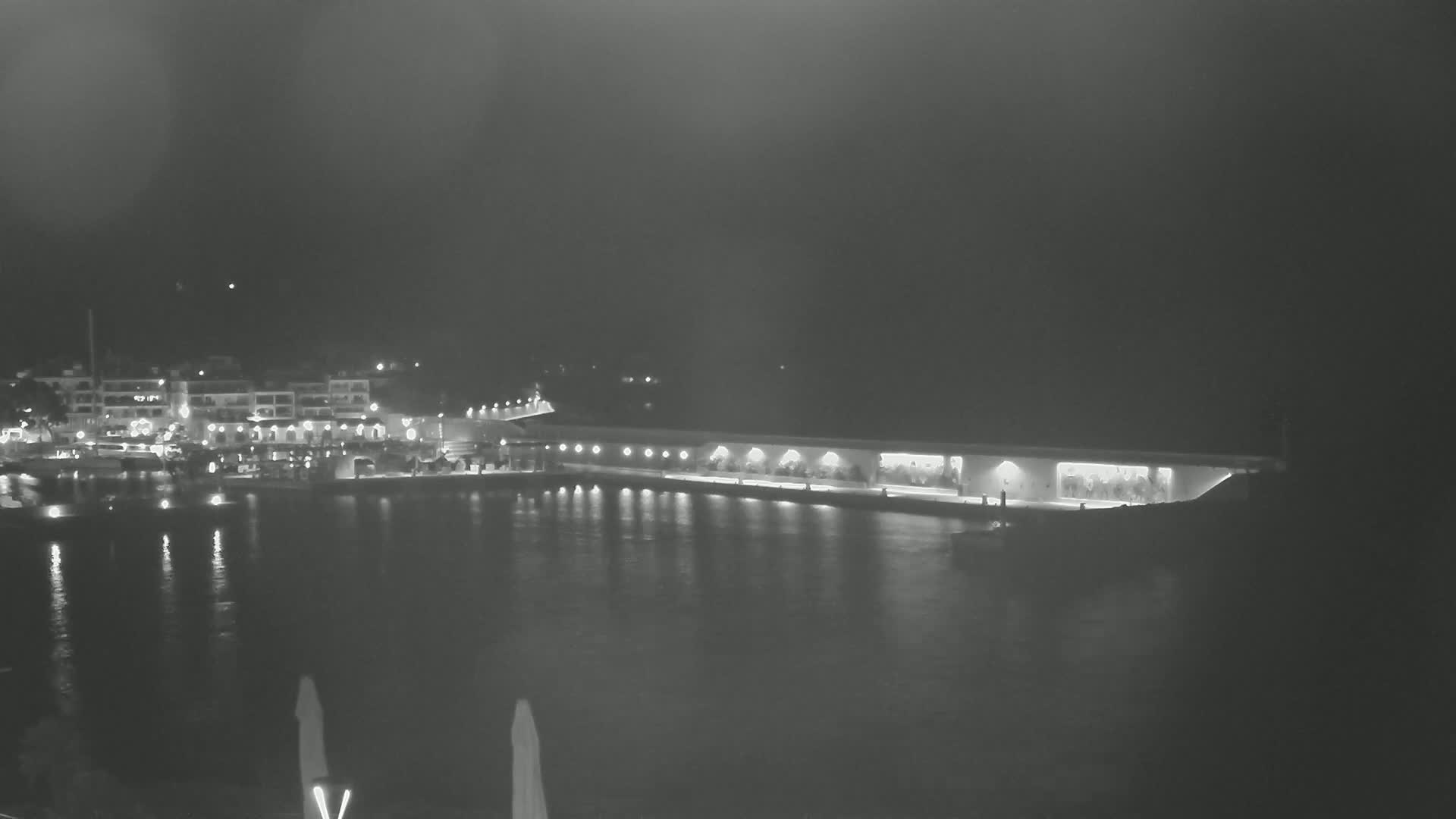 A nighttime view of a harbor with boats docked, illuminated buildings along the shore, and a long, lit structure extending into the calm water.