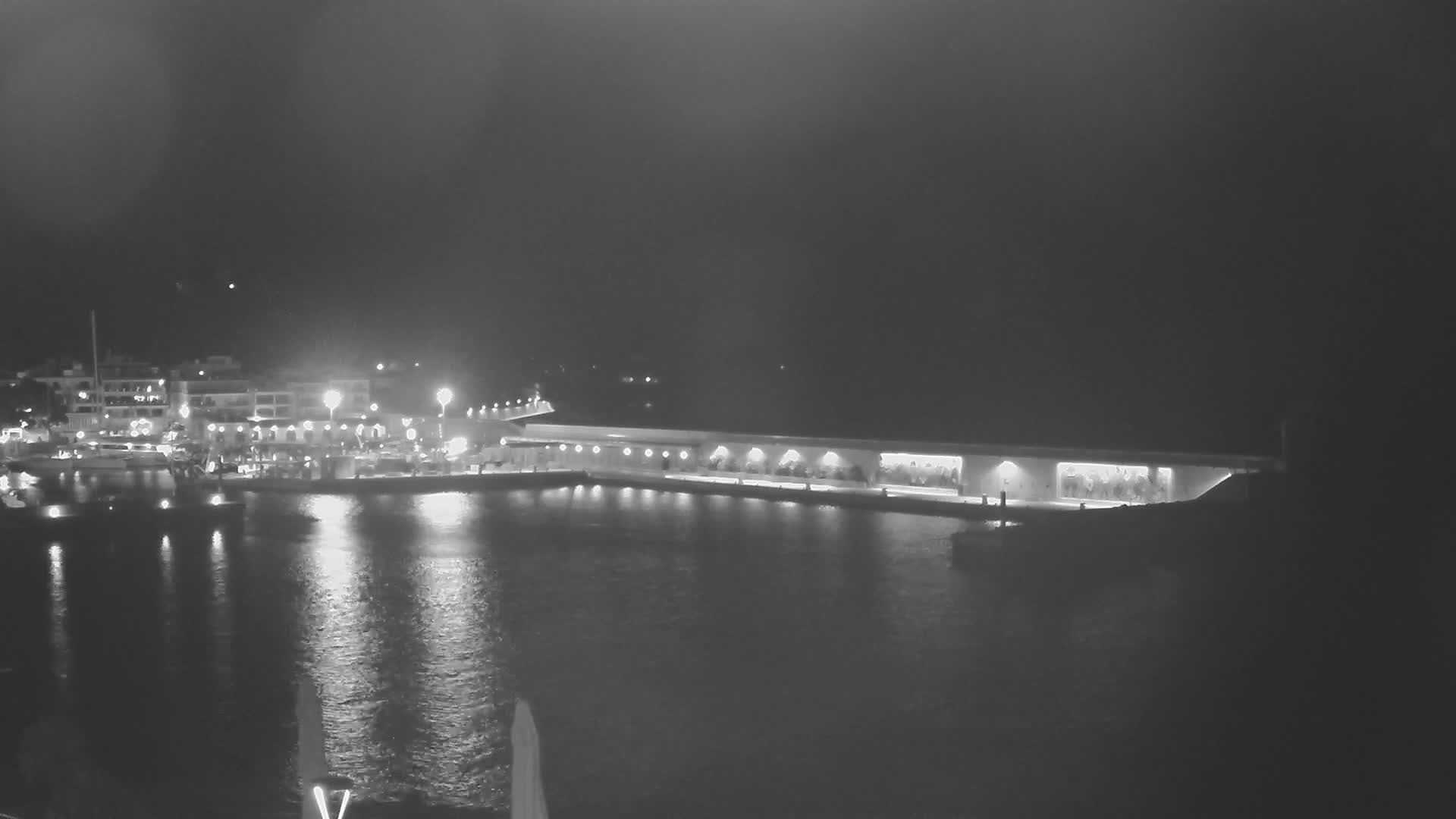 A nighttime harbor scene shows a long, low building with lit windows along a calm body of water, opposite a town with lights reflecting on the water.
