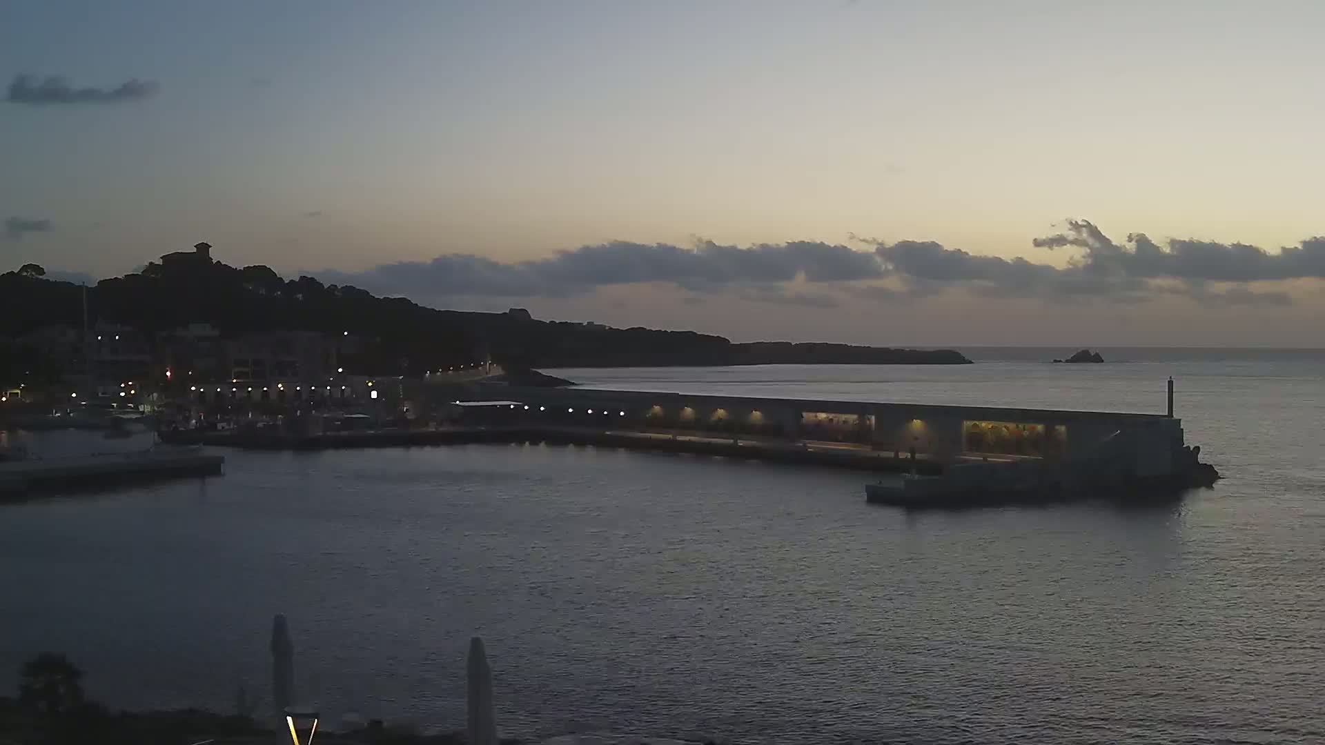 A harbor at twilight with a long pier extending into calm water, backed by a hilltop town and a dark coastline under a cloudy sky.