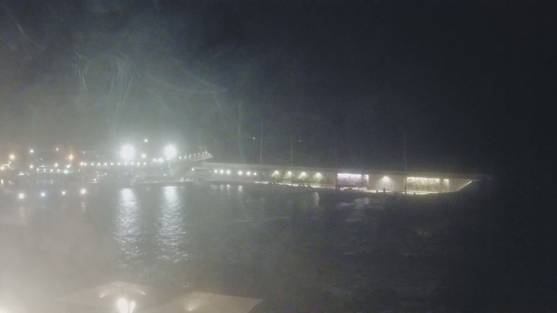 A hazy night scene shows a brightly lit waterfront area with boats moored alongside a long, low building.