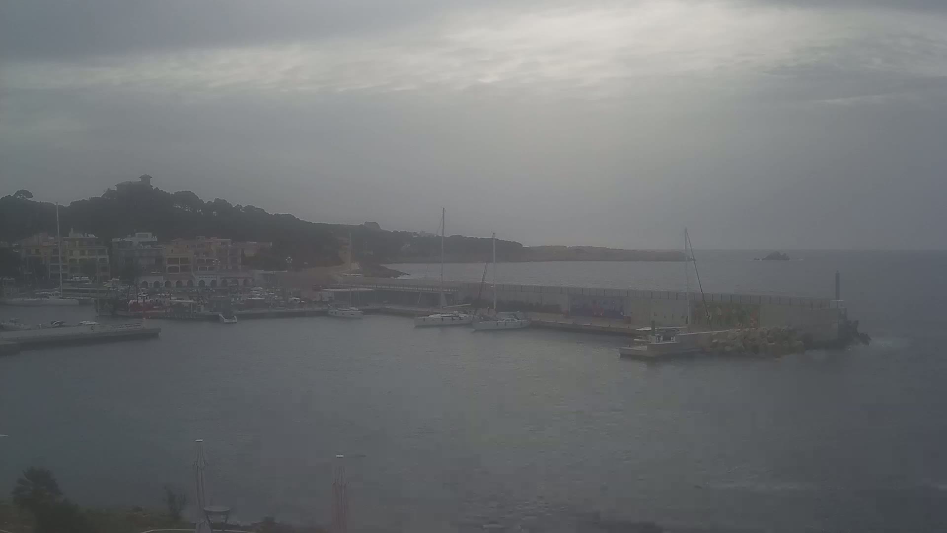 A hazy, overcast day reveals a harbor with several boats, a stone seawall, and buildings on a nearby hillside.